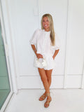 Woman in a white top and shorts set with gold sandals holding a white purse and standing against a white wall.