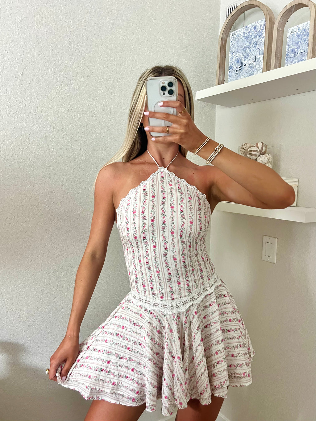 Woman taking a mirror selfie wearing a white and pink floral halter dress in a room with a shelf and decorative items
