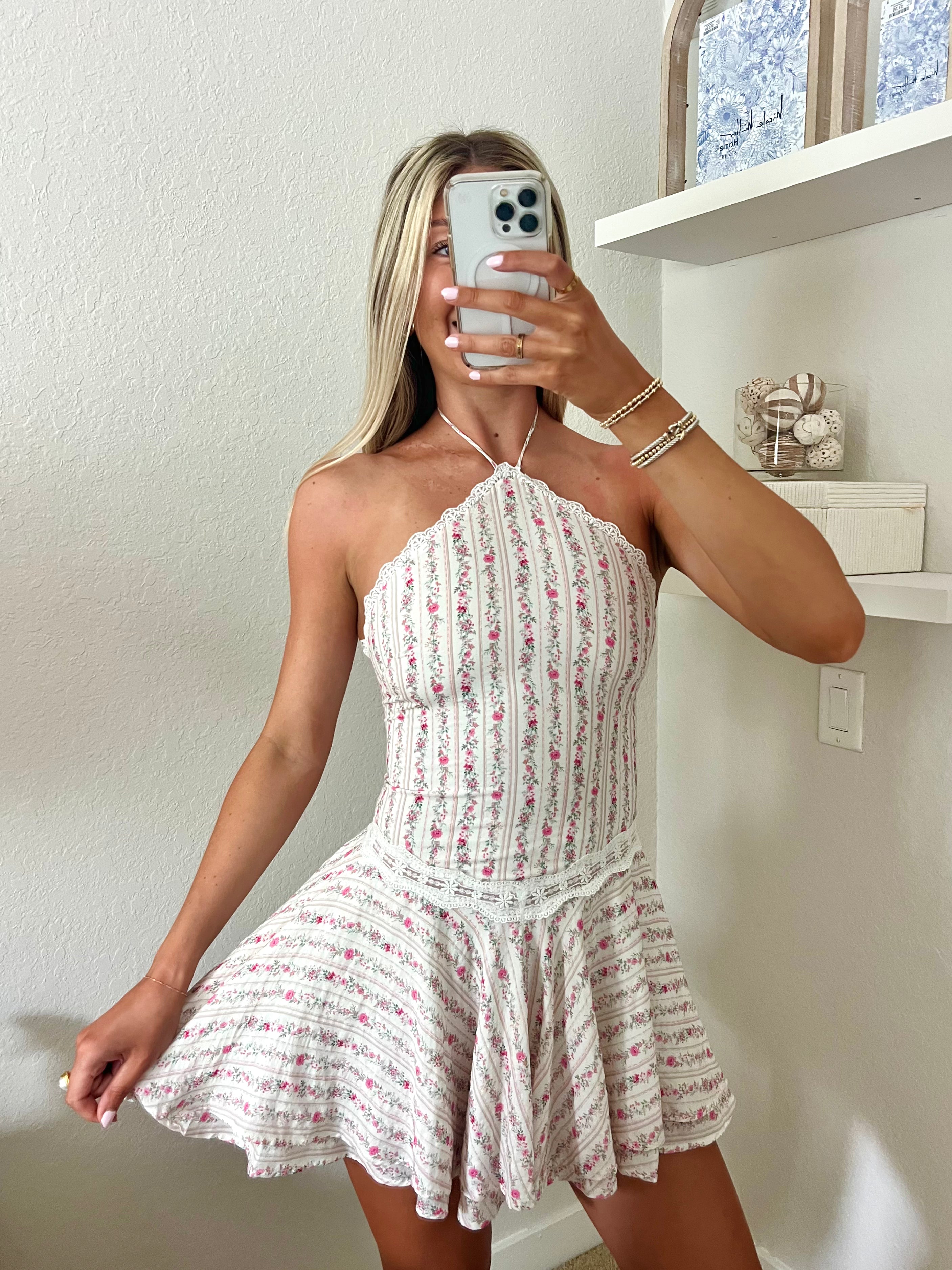 Woman taking a mirror selfie wearing a white and pink floral halter dress in a room with a shelf and decorative items