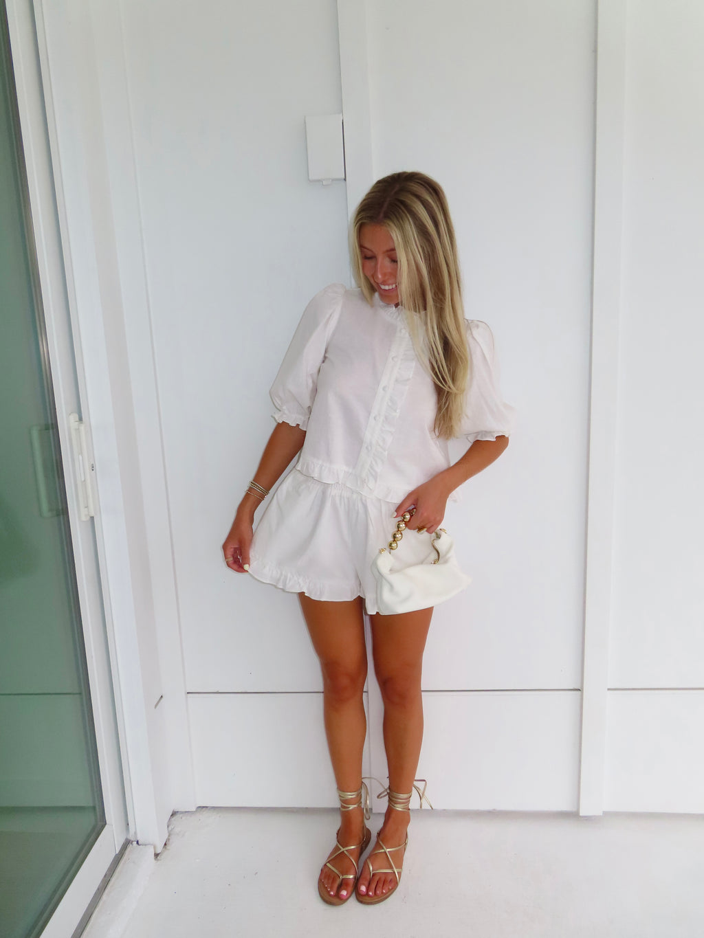 Woman in a white top and shorts set with gold sandals holding a white purse and standing against a white wall.