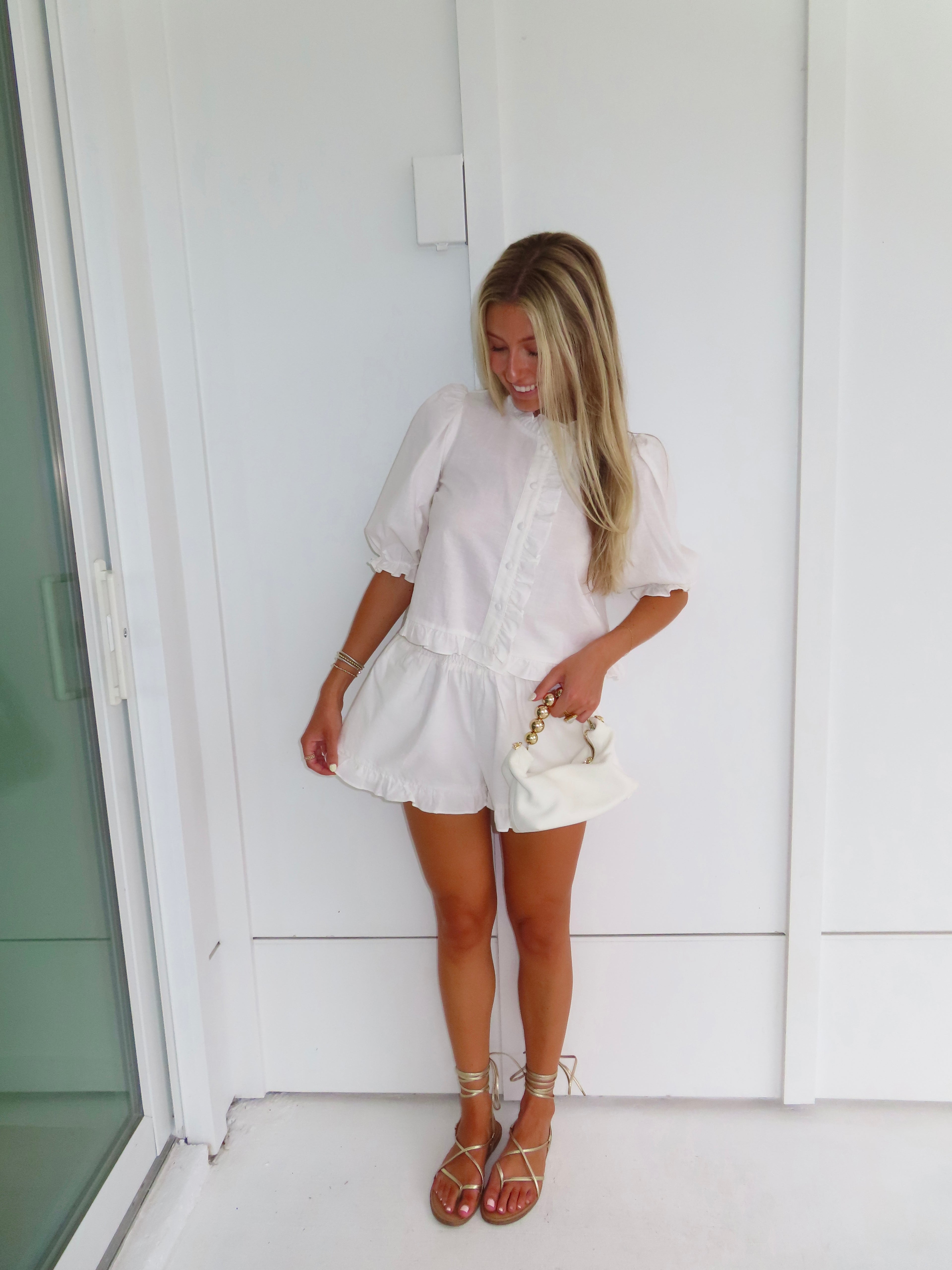 Woman in a white top and shorts set with gold sandals holding a white purse and standing against a white wall.