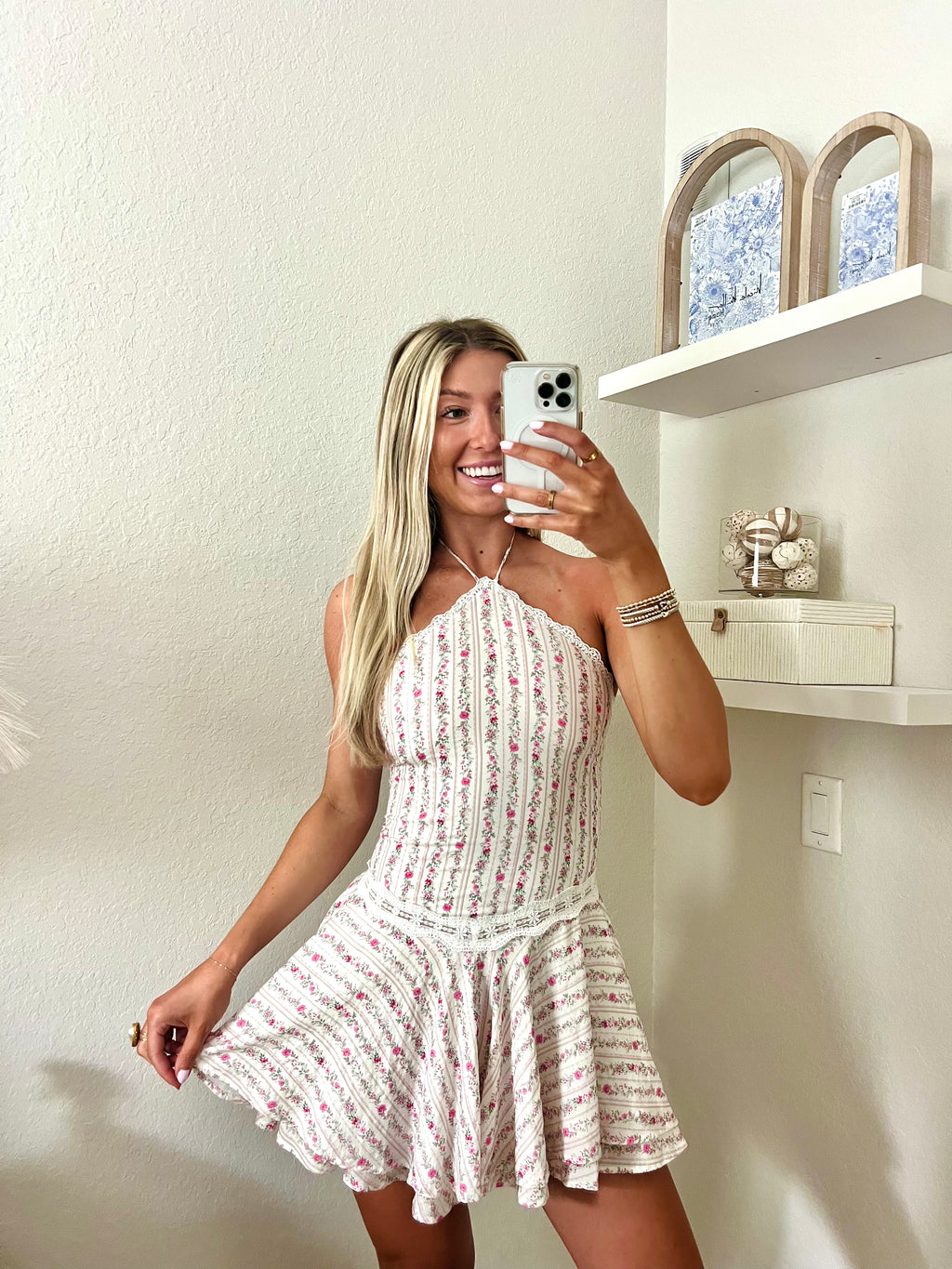 Woman taking a mirror selfie wearing a white and pink floral halter dress in a room with a shelf and decorative items