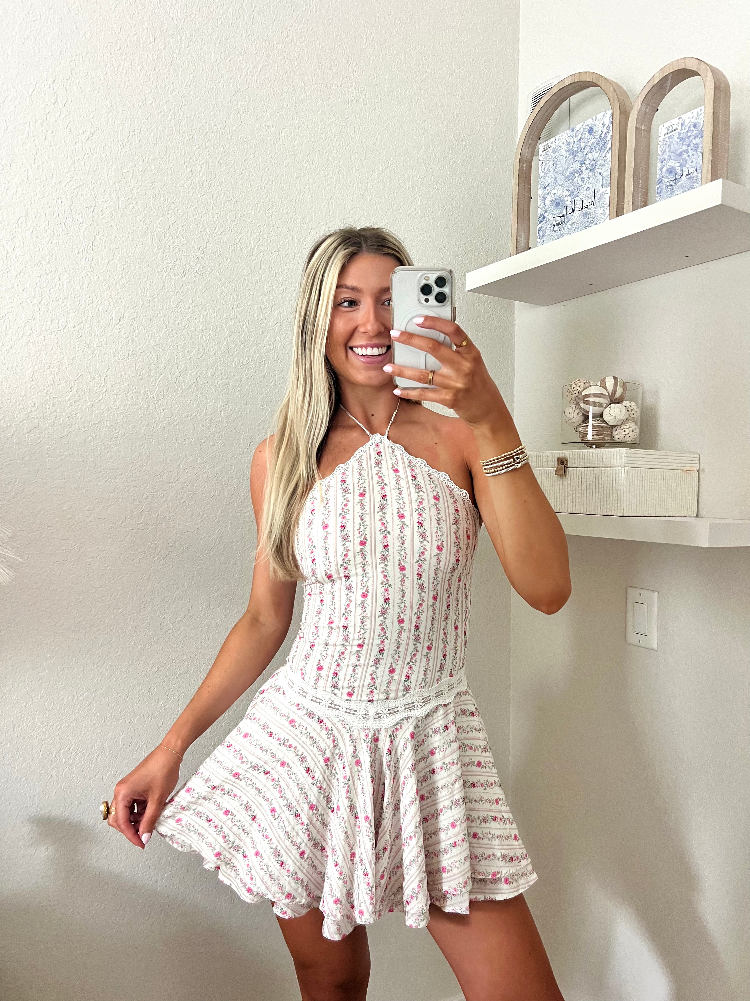 Woman taking a mirror selfie wearing a white and pink floral halter dress in a room with a shelf and decorative items
