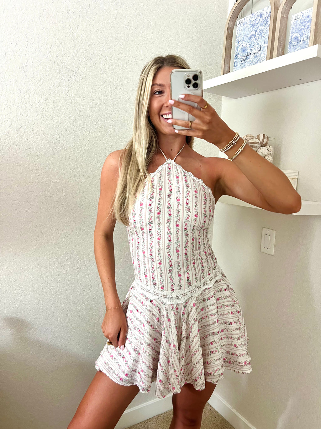 Woman taking a mirror selfie wearing a white and pink floral halter dress in a room with a shelf and decorative items