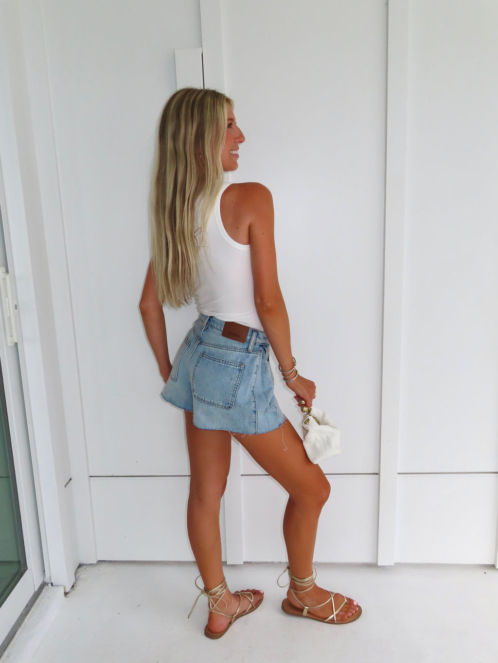 Woman in white tank top and denim shorts holding a white purse standing against a white wall.