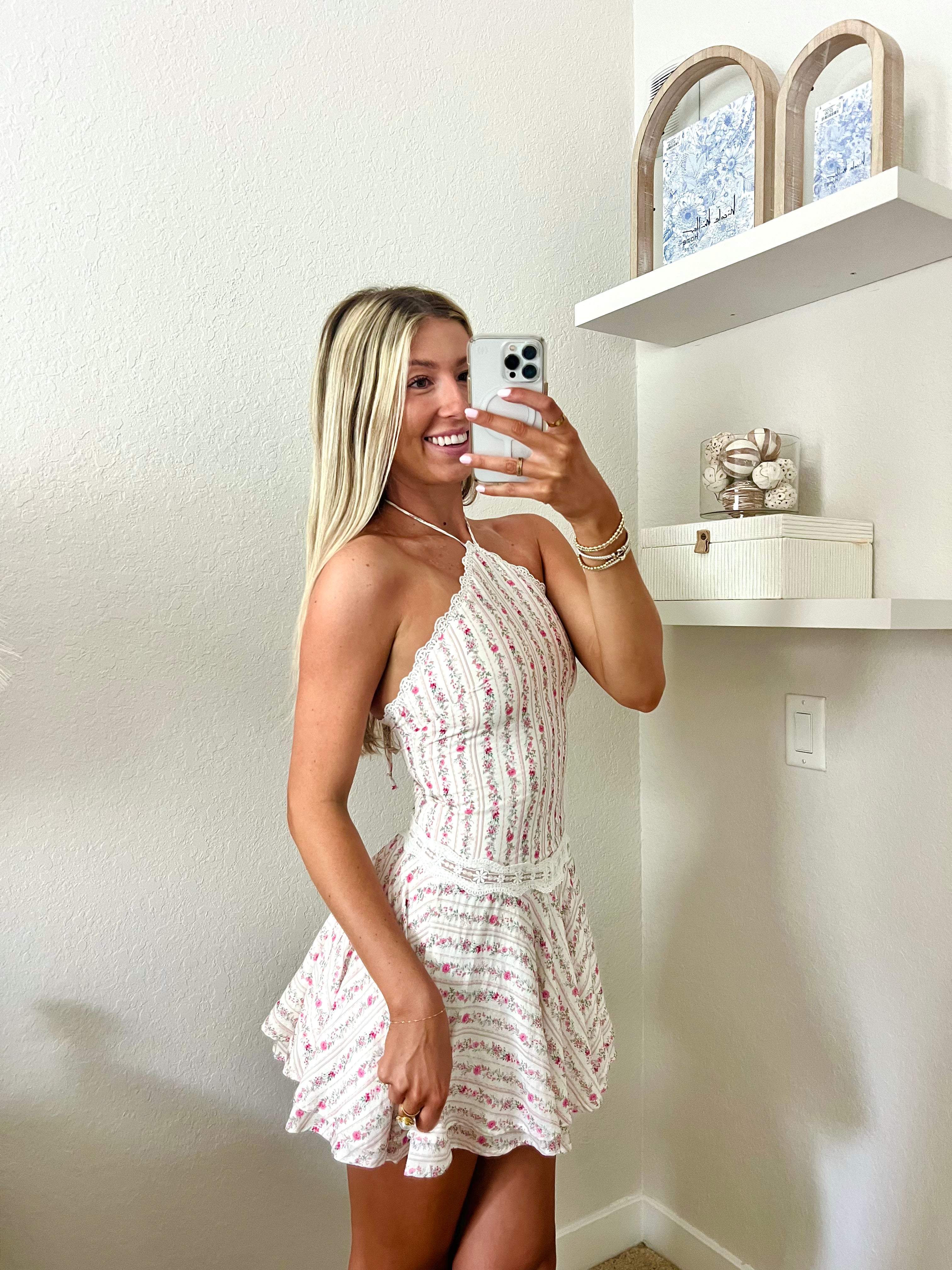 Woman taking a mirror selfie wearing a white and pink floral halter dress in a room with a shelf and decorative items