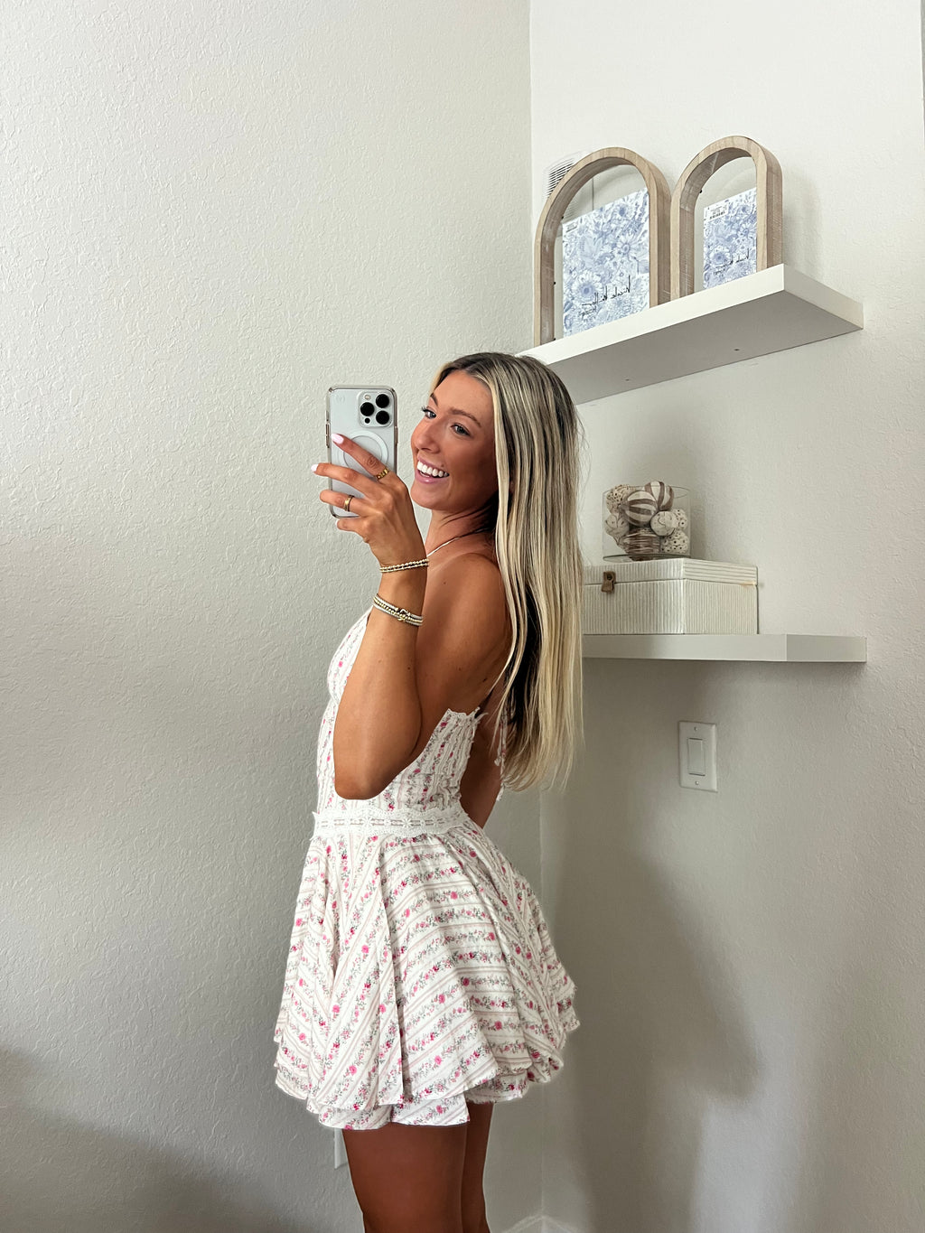 Woman taking a mirror selfie wearing a white and pink floral halter dress in a room with a shelf and decorative items