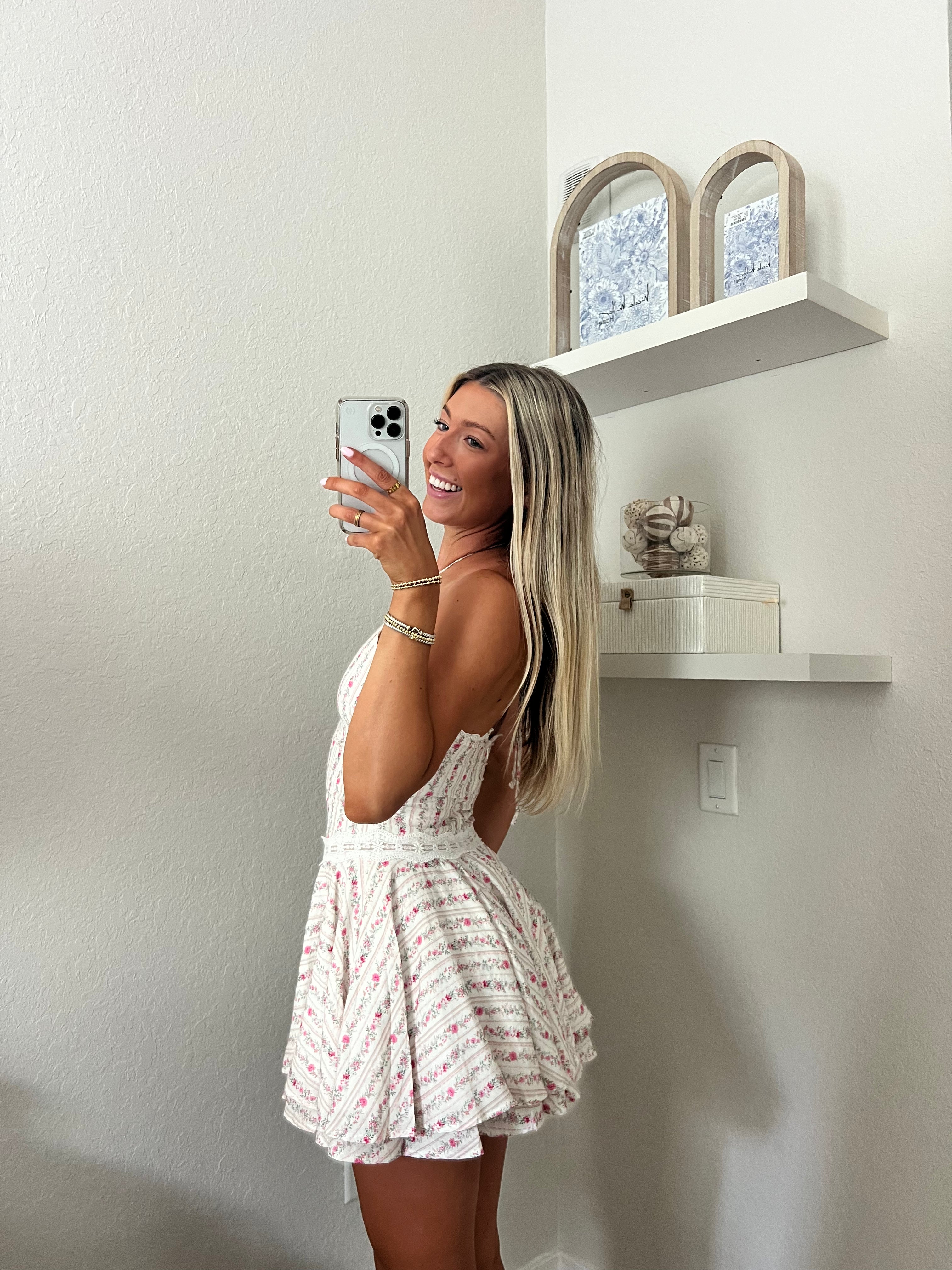 Woman taking a mirror selfie wearing a white and pink floral halter dress in a room with a shelf and decorative items