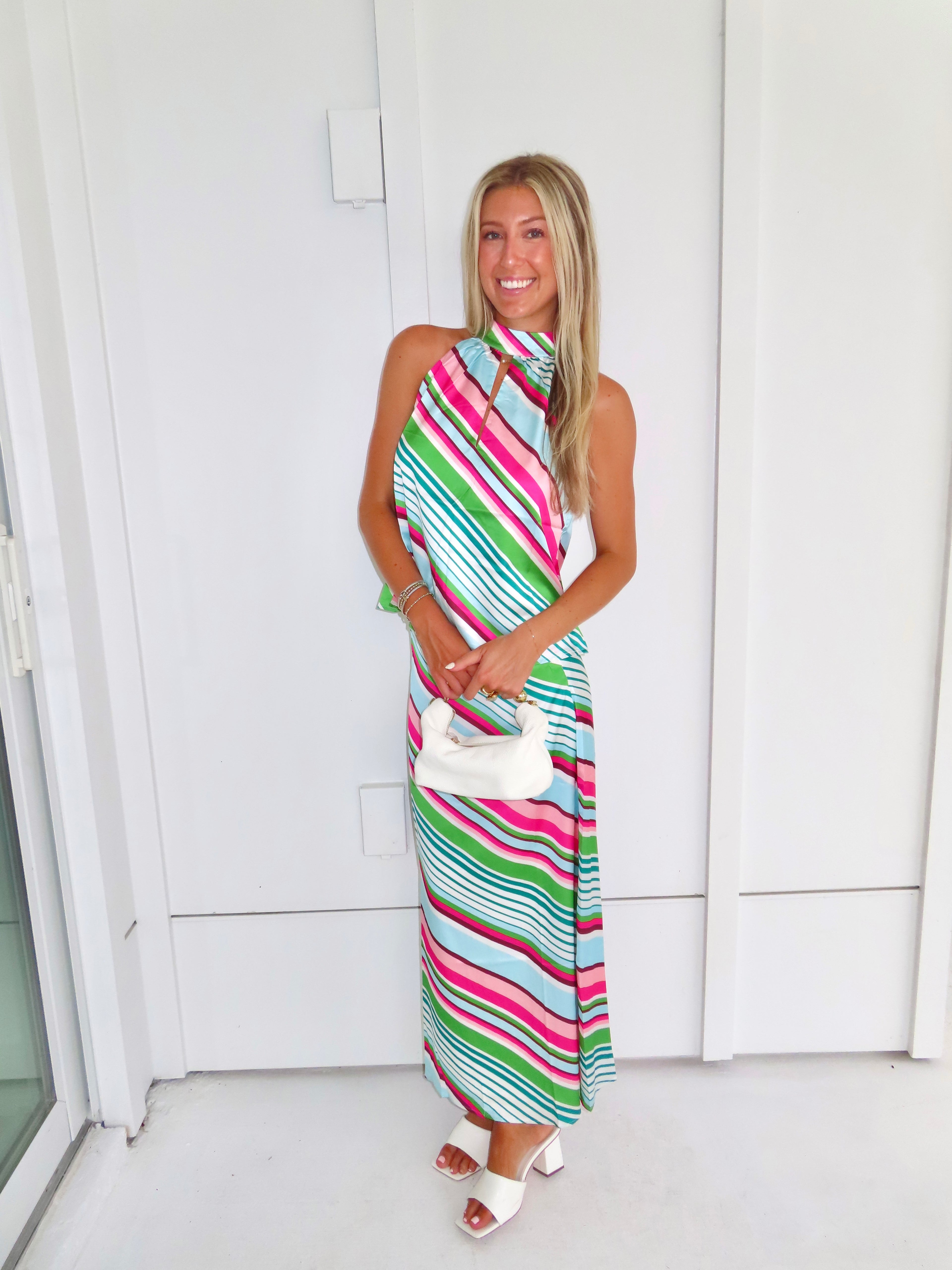 Woman wearing a colorful striped maxi skirt set holding a white handbag and standing in front of a white wall.