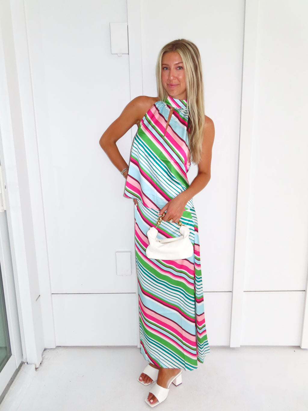 Woman wearing a colorful striped maxi skirt set holding a white handbag and standing in front of a white wall.