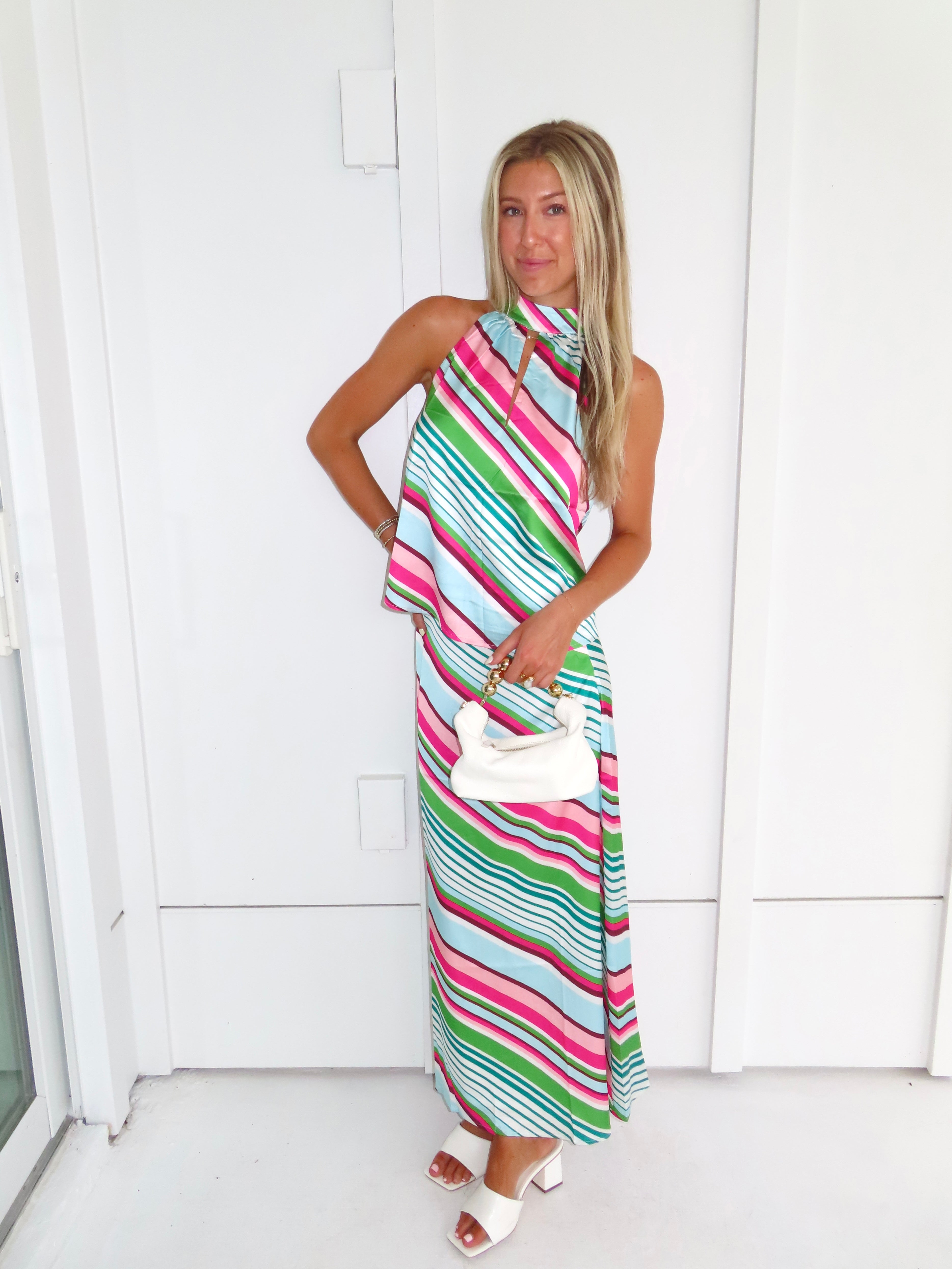 Woman wearing a colorful striped maxi skirt set holding a white handbag and standing in front of a white wall.