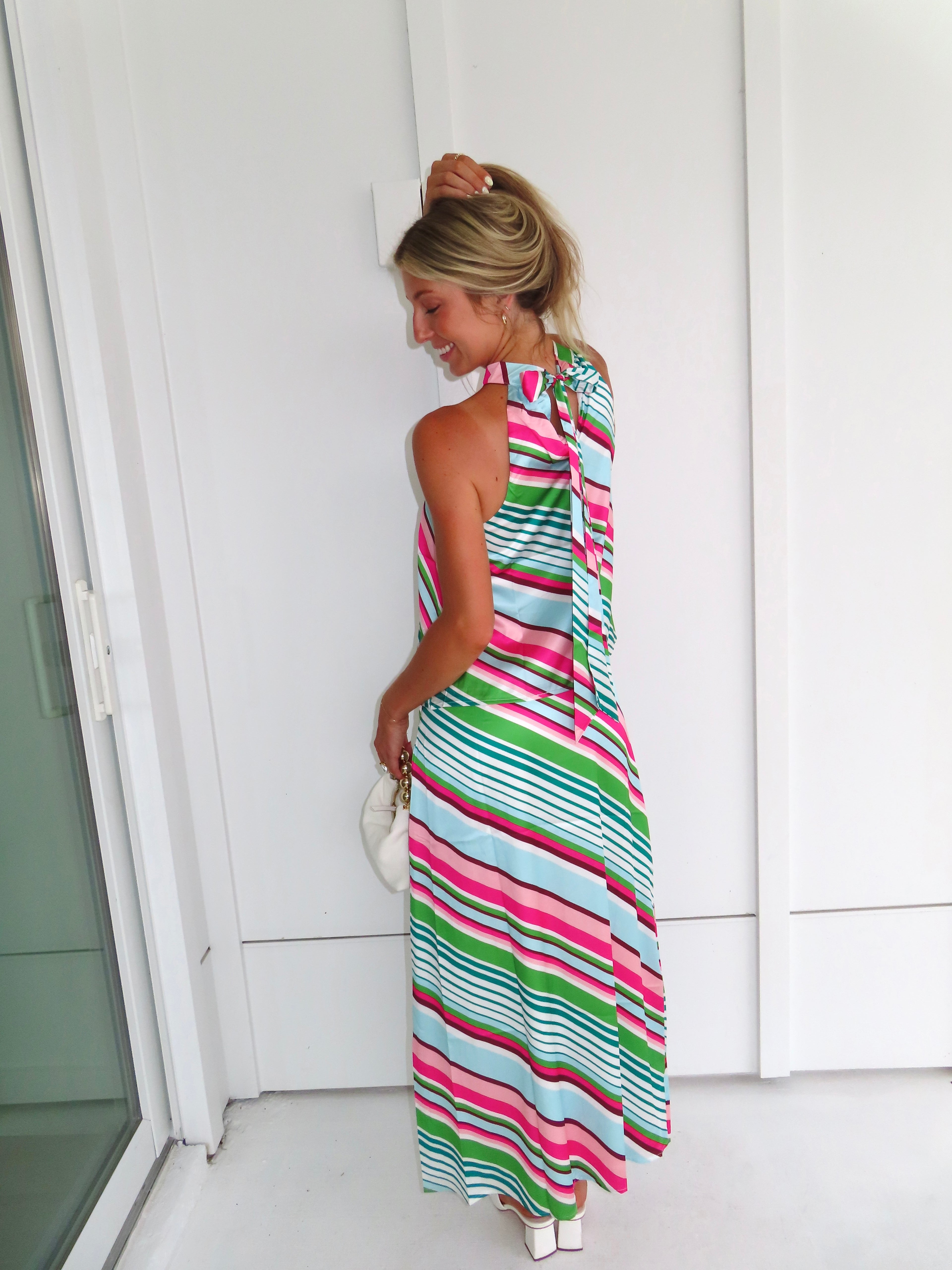 Woman wearing a colorful striped maxi skirt set holding a white handbag and standing in front of a white wall.