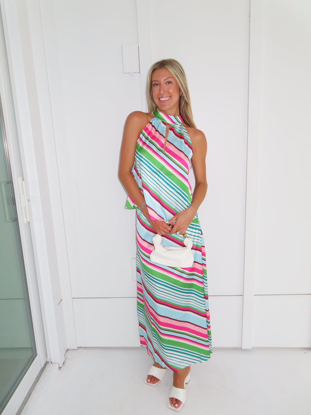 Woman wearing a colorful striped maxi skirt set holding a white handbag and standing in front of a white wall.