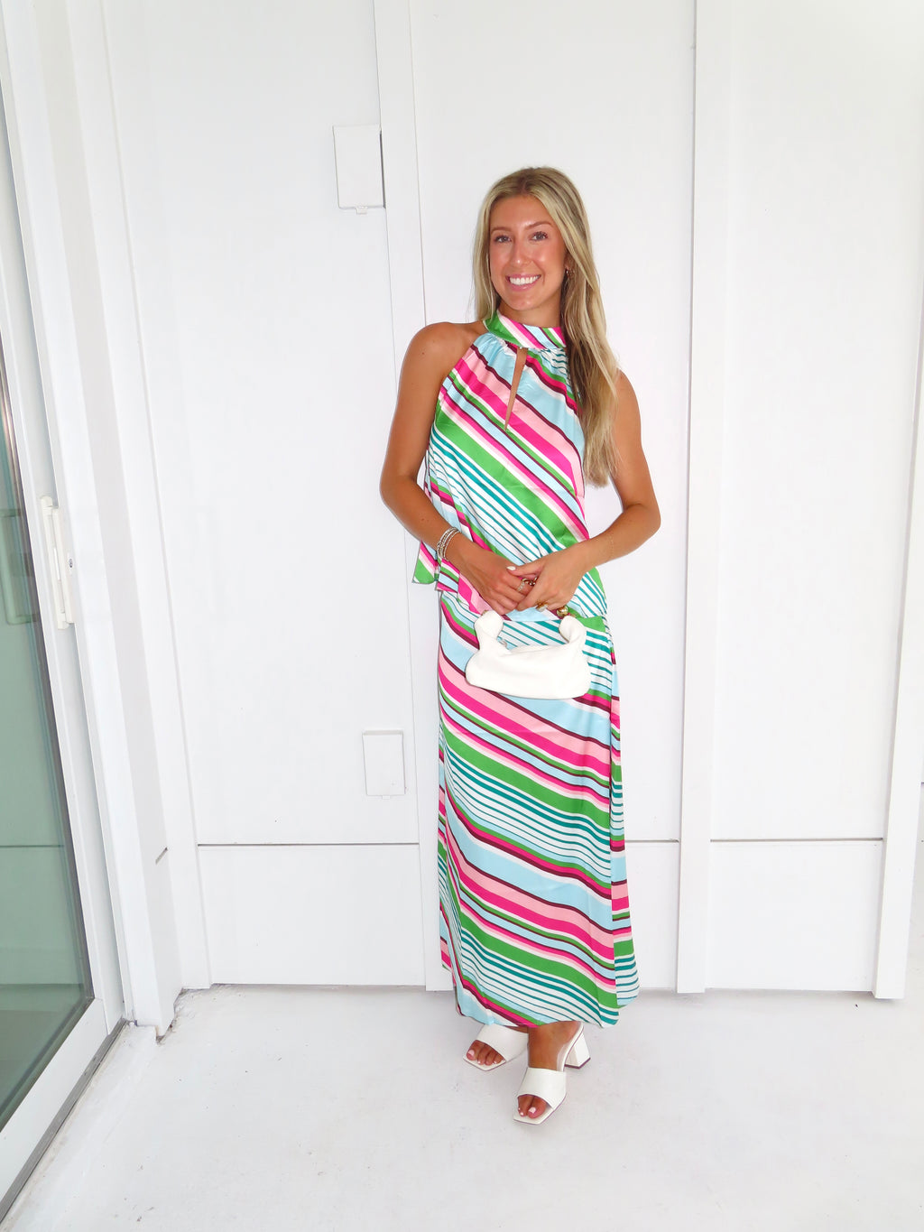 Woman wearing a colorful striped maxi skirt set holding a white handbag and standing in front of a white wall.