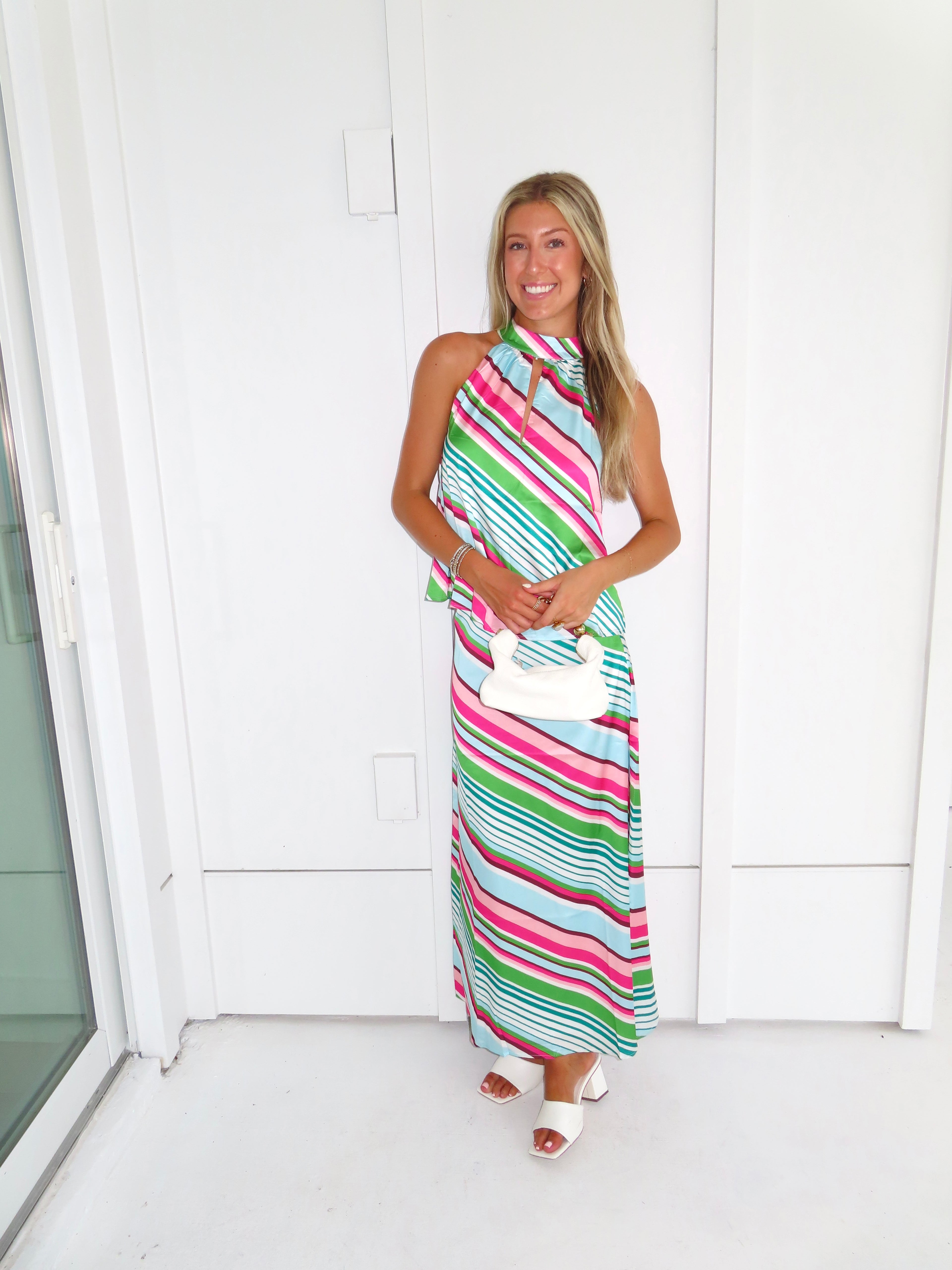 Woman wearing a colorful striped maxi skirt set holding a white handbag and standing in front of a white wall.