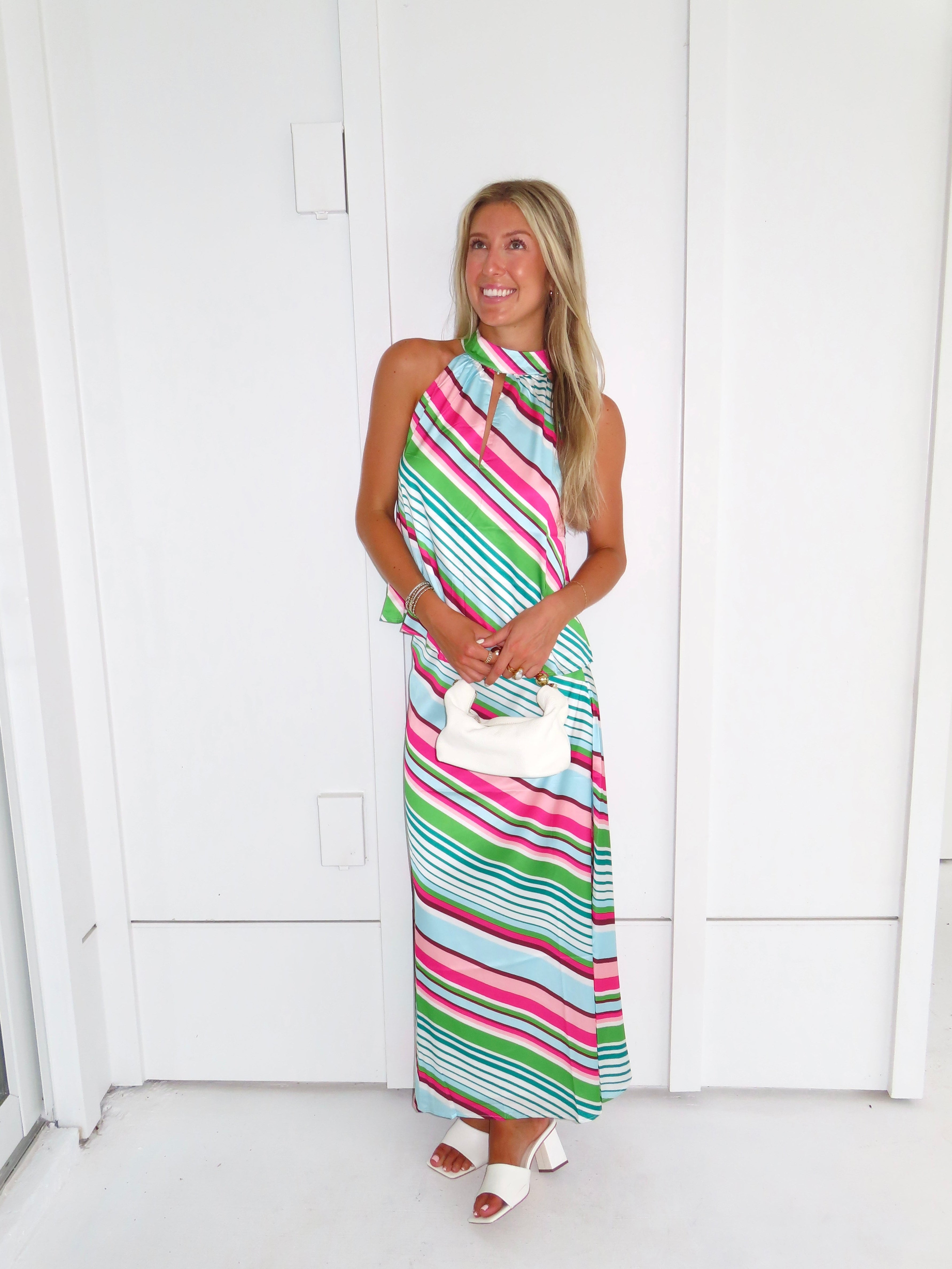 Woman wearing a colorful striped maxi skirt set holding a white handbag and standing in front of a white wall.