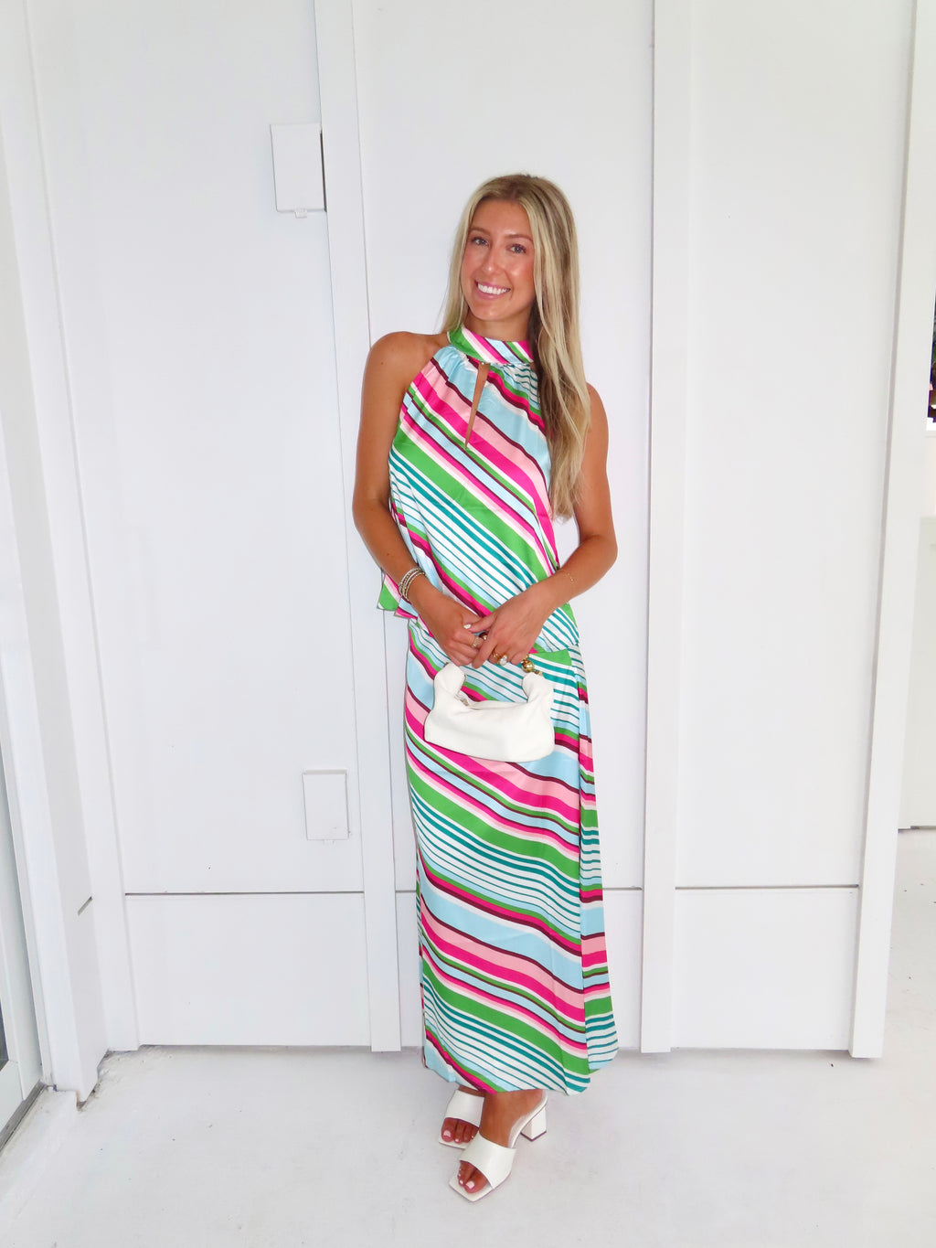 Woman wearing a colorful striped maxi skirt set holding a white handbag and standing in front of a white wall.