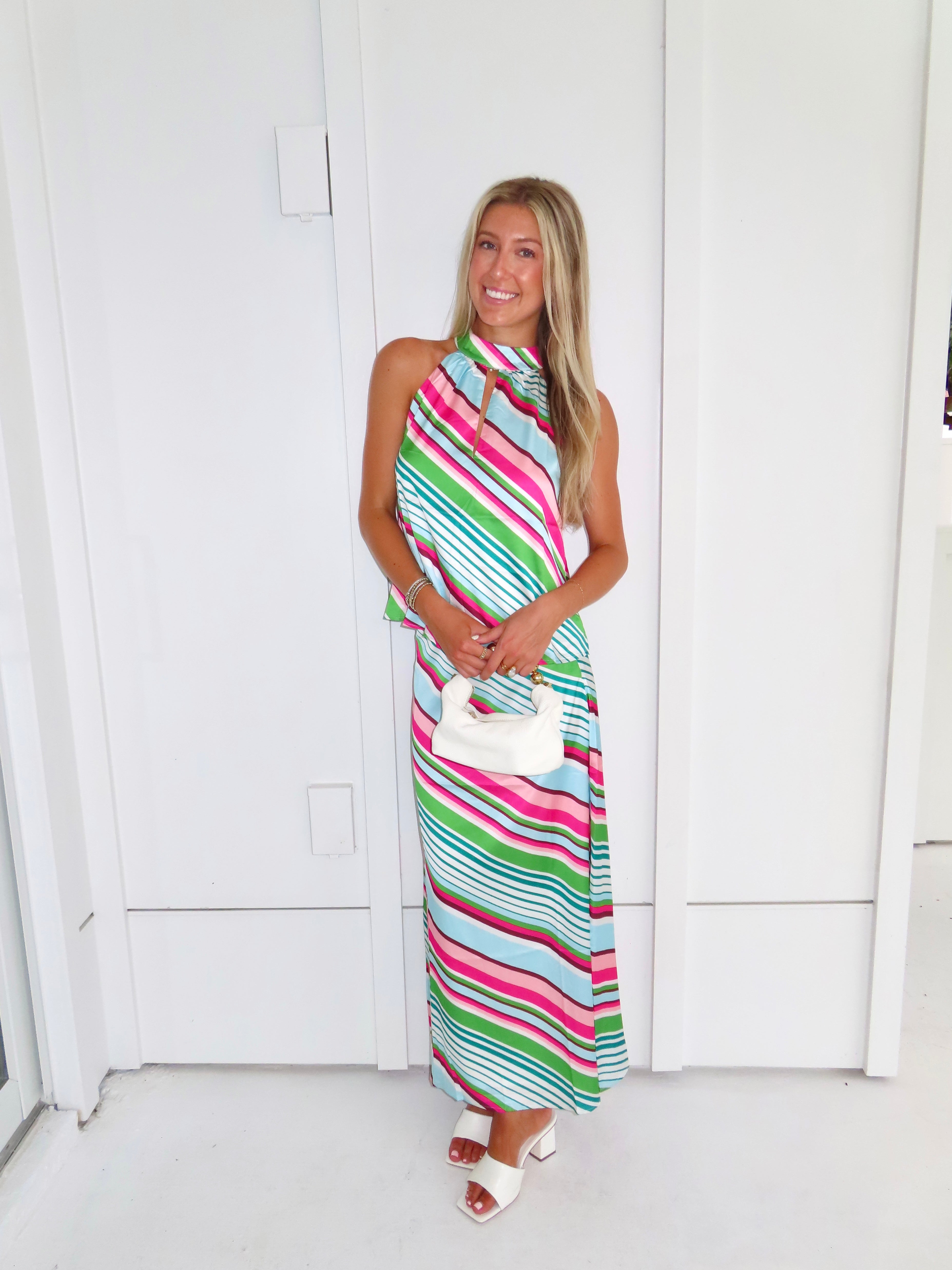Woman wearing a colorful striped maxi skirt set holding a white handbag and standing in front of a white wall.