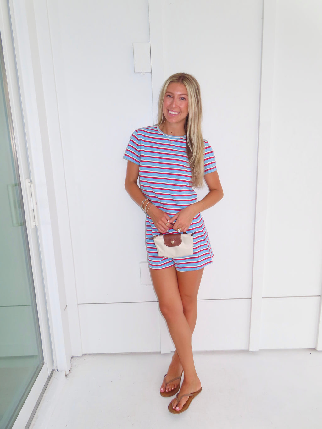 Woman wearing a red, white, and blue striped shirt and short set holding a cream purse and standing against a white wall.