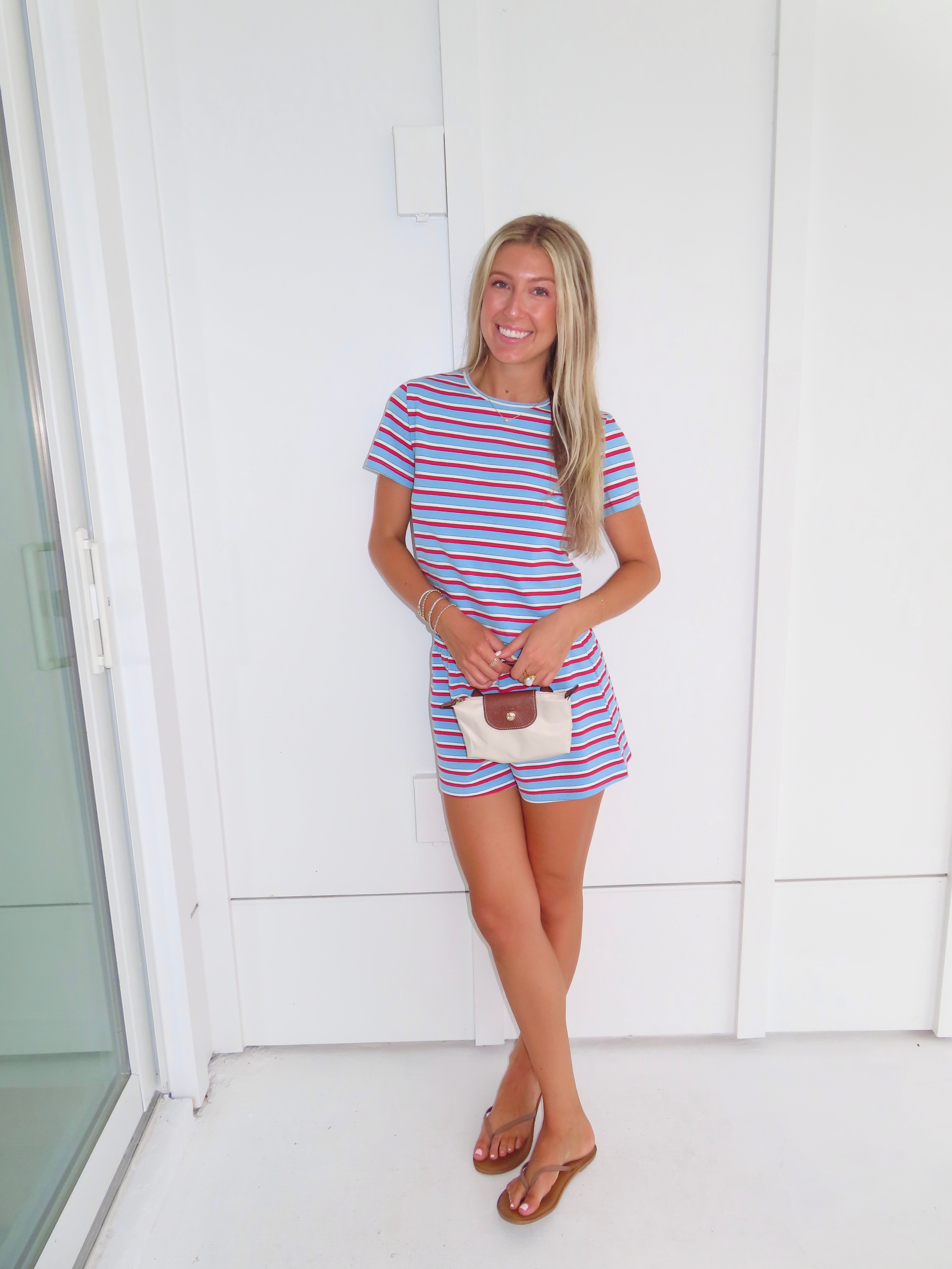 Woman wearing a red, white, and blue striped shirt and short set holding a cream purse and standing against a white wall.