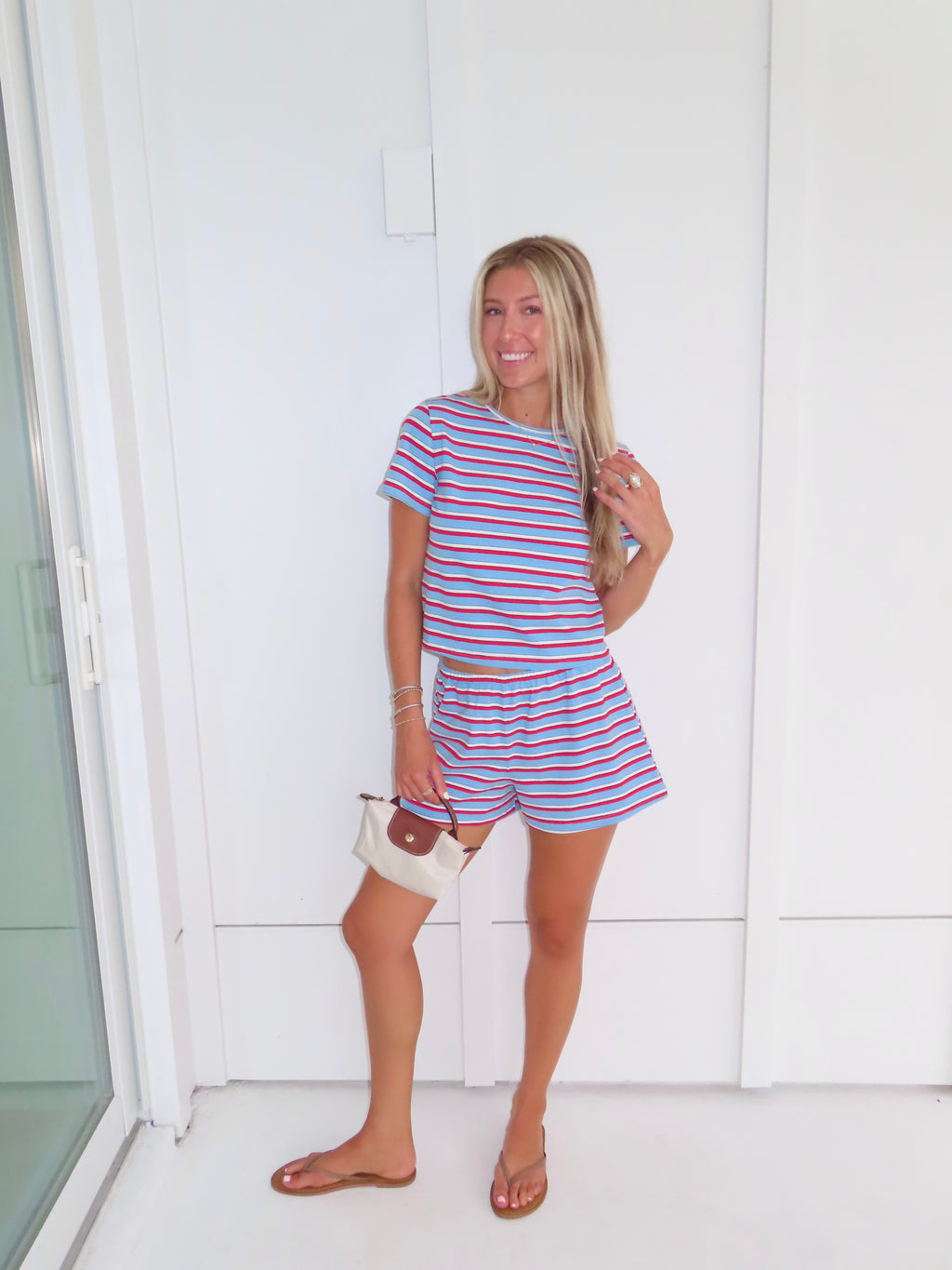 Woman wearing a red, white, and blue striped shirt and short set holding a cream purse and standing against a white wall.