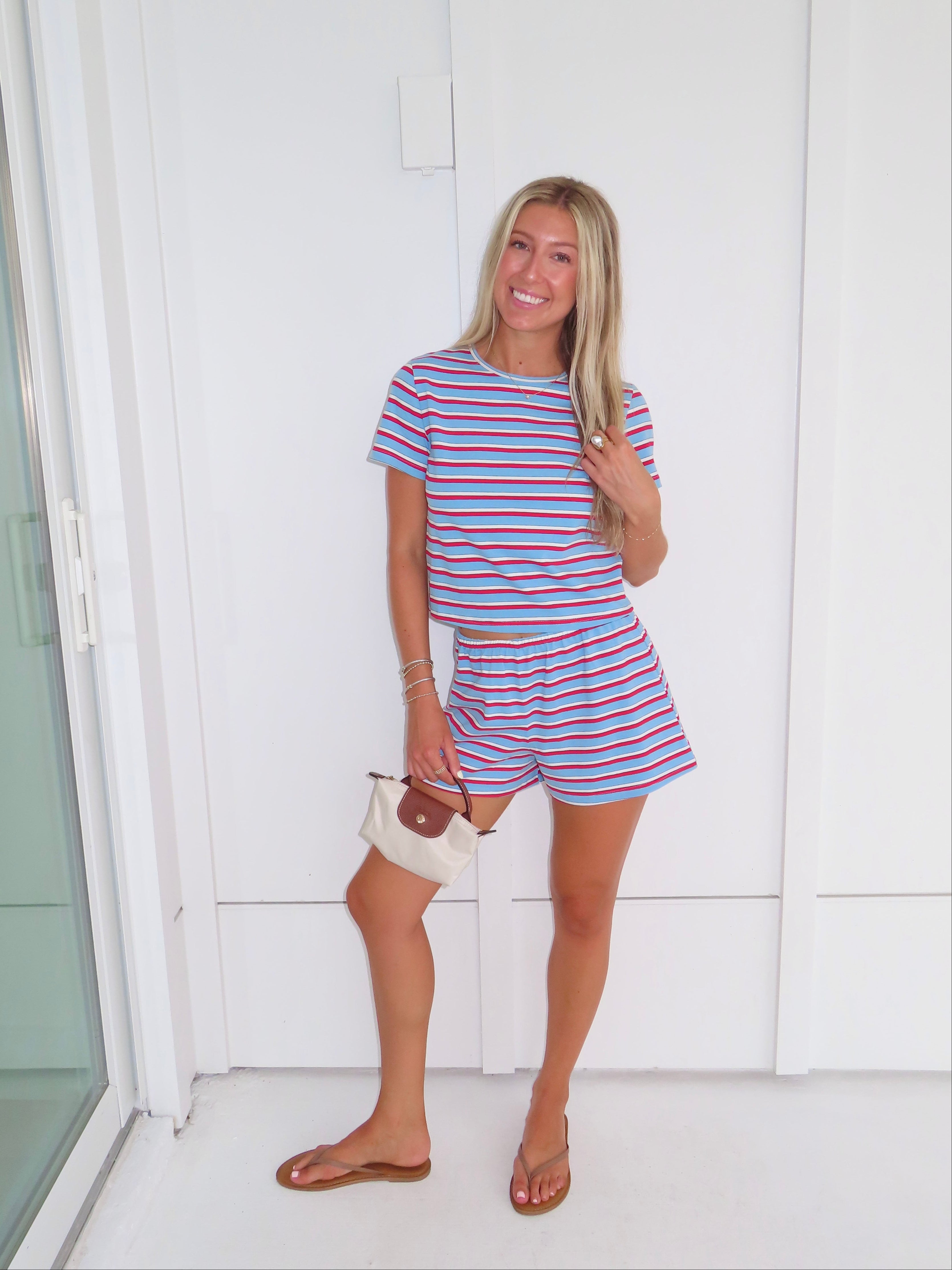 Woman wearing a red, white, and blue striped shirt and short set holding a cream purse and standing against a white wall.