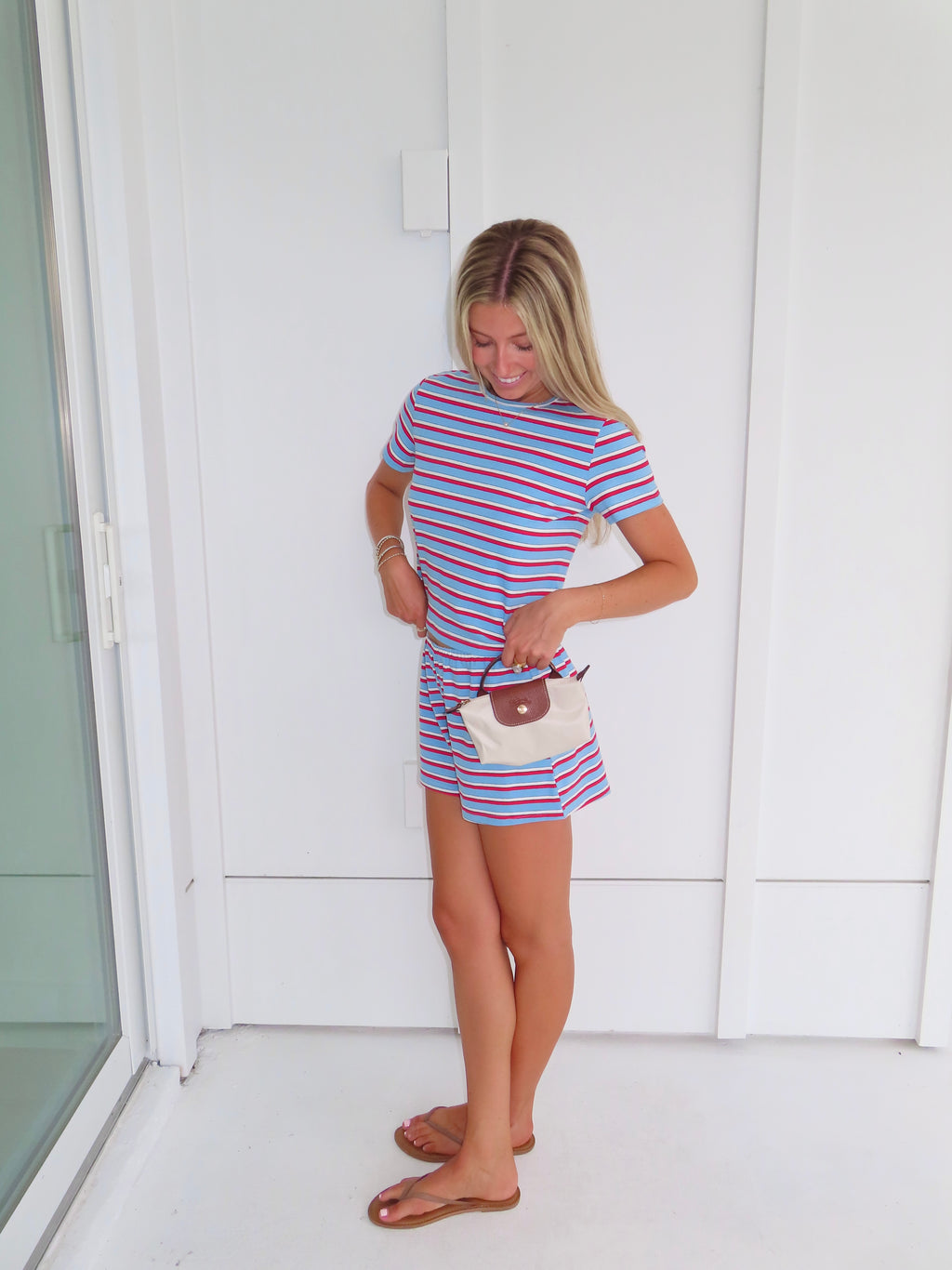 Woman wearing a red, white, and blue striped shirt and short set holding a cream purse and standing against a white wall.