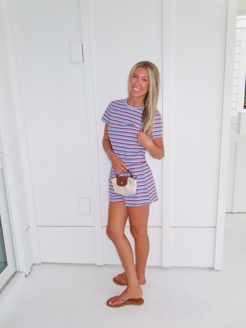 Woman wearing a red, white, and blue striped shirt and short set holding a cream purse and standing against a white wall.