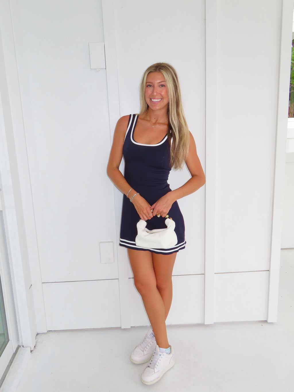 Woman wearing a navy dress with white trim and white sneakers holding a white purse against a white wall.