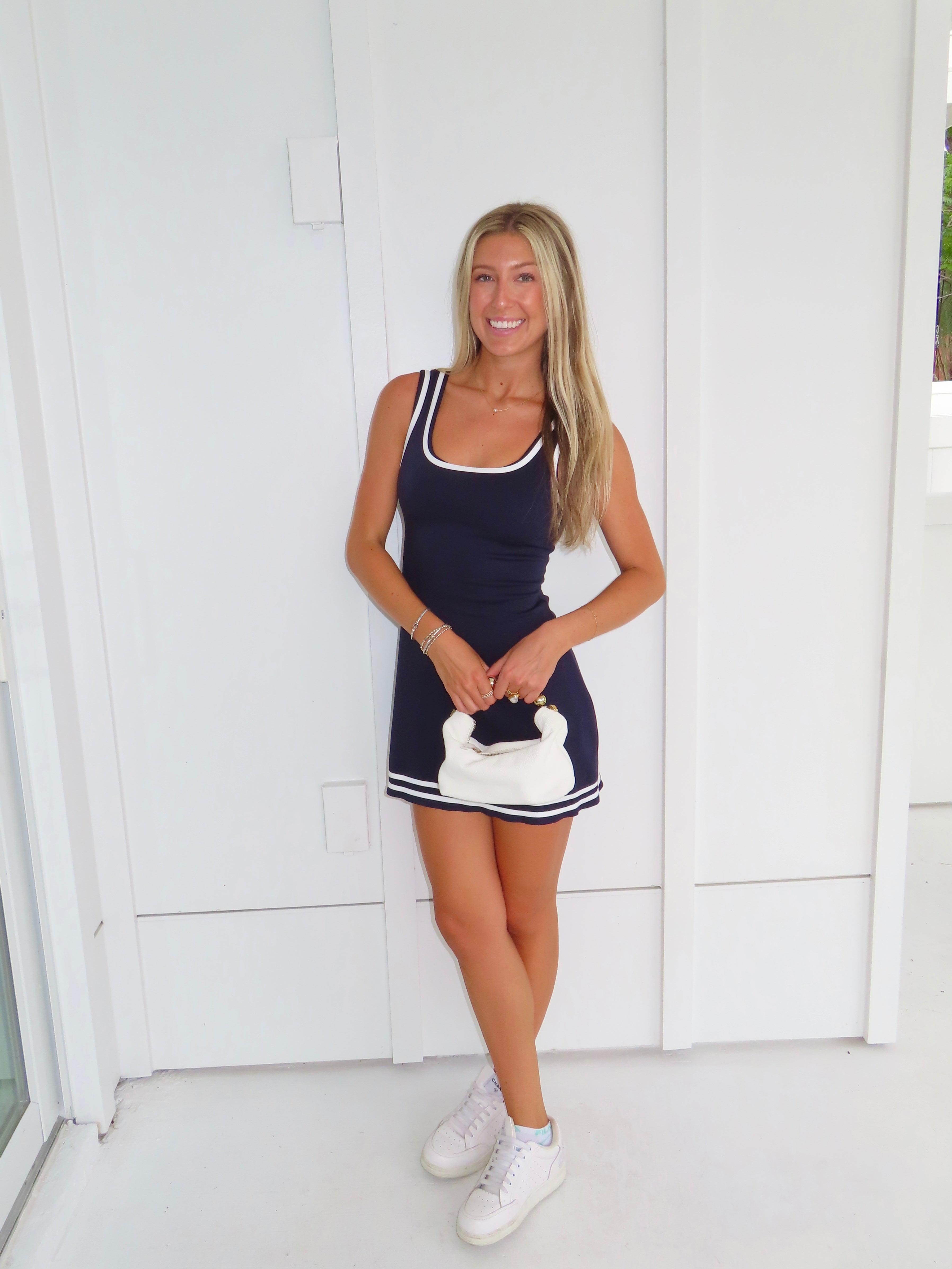 Woman wearing a navy dress with white trim and white sneakers holding a white purse against a white wall.