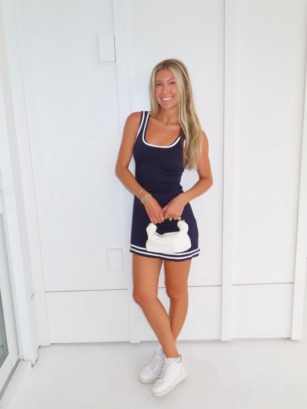 Woman wearing a navy dress with white trim and white sneakers holding a white purse against a white wall.