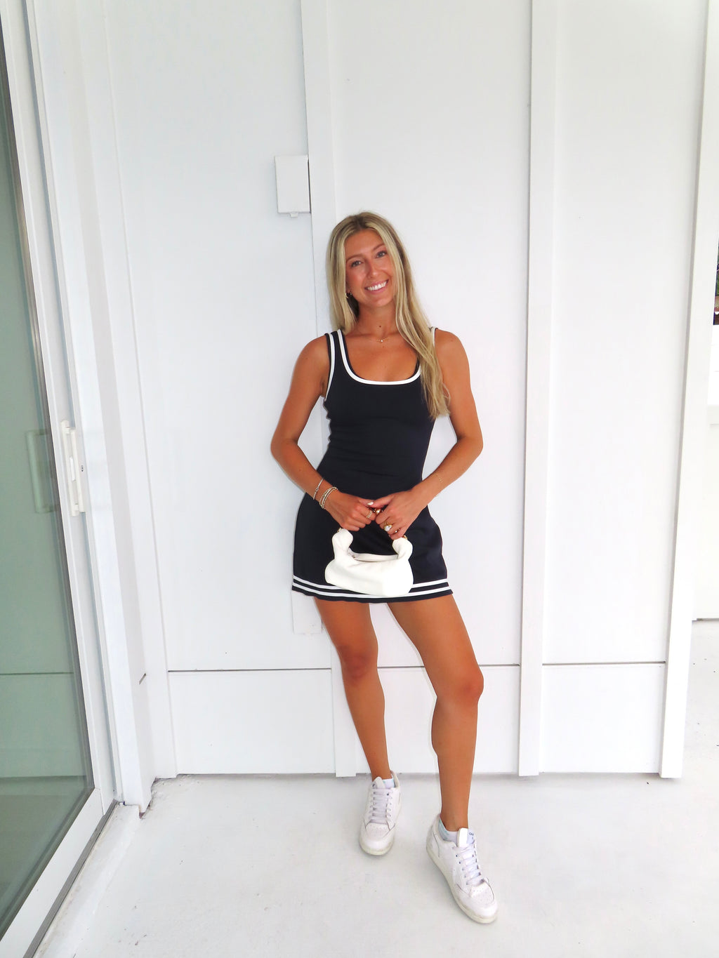 Woman wearing a navy dress with white trim and white sneakers holding a white purse against a white wall.