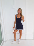 Woman wearing a navy dress with white trim and white sneakers holding a white purse against a white wall.