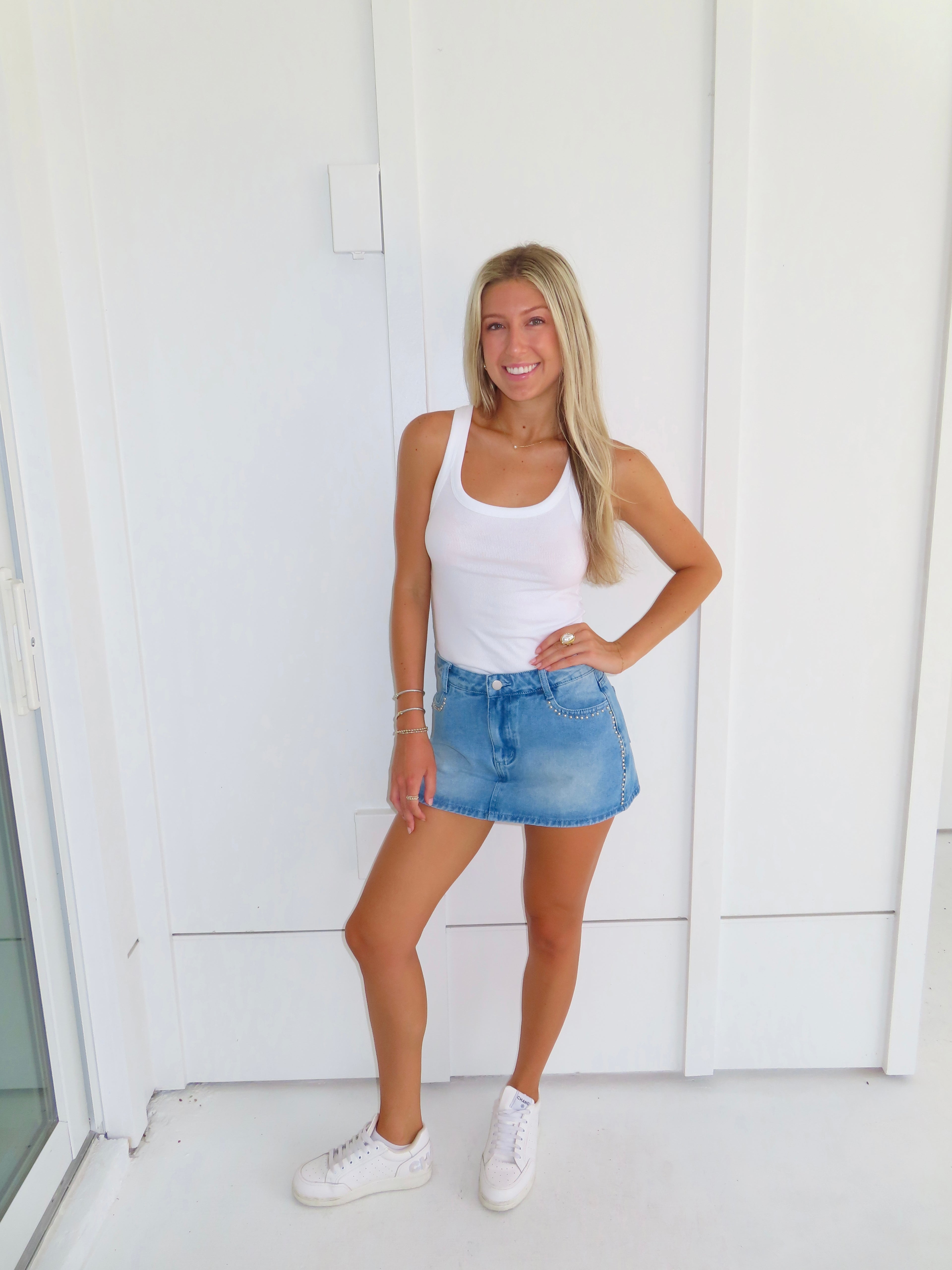 Woman wearing a white tank top and denim studded skirt with white sneakers standing against a white wall.