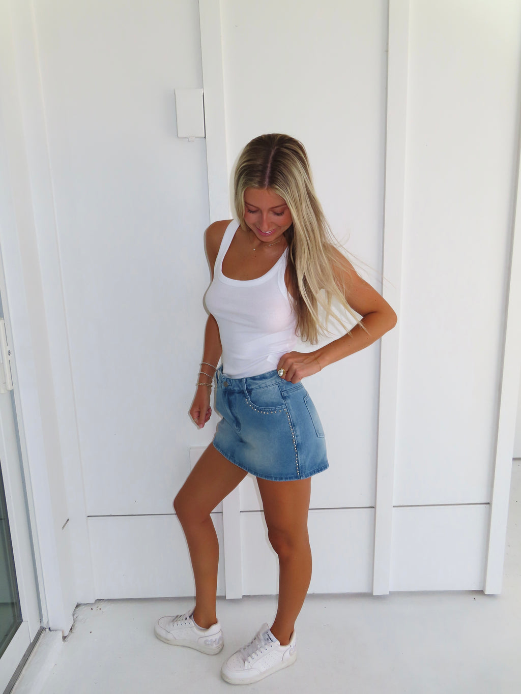 Woman wearing a white tank top and denim studded skirt with white sneakers standing against a white wall.