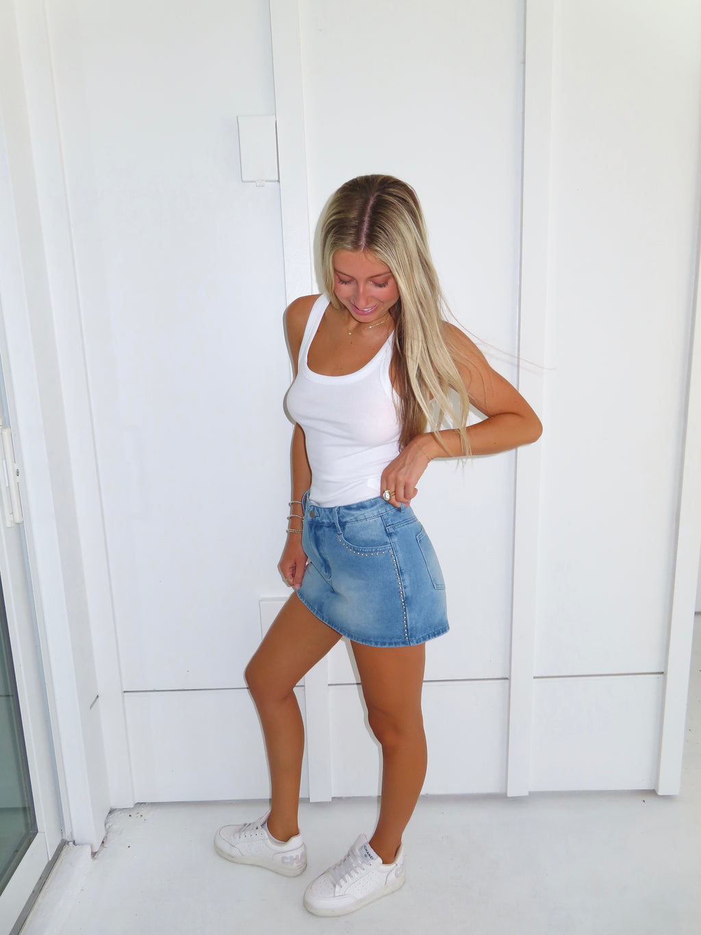 Woman wearing a white tank top and denim studded skirt with white sneakers standing against a white wall.