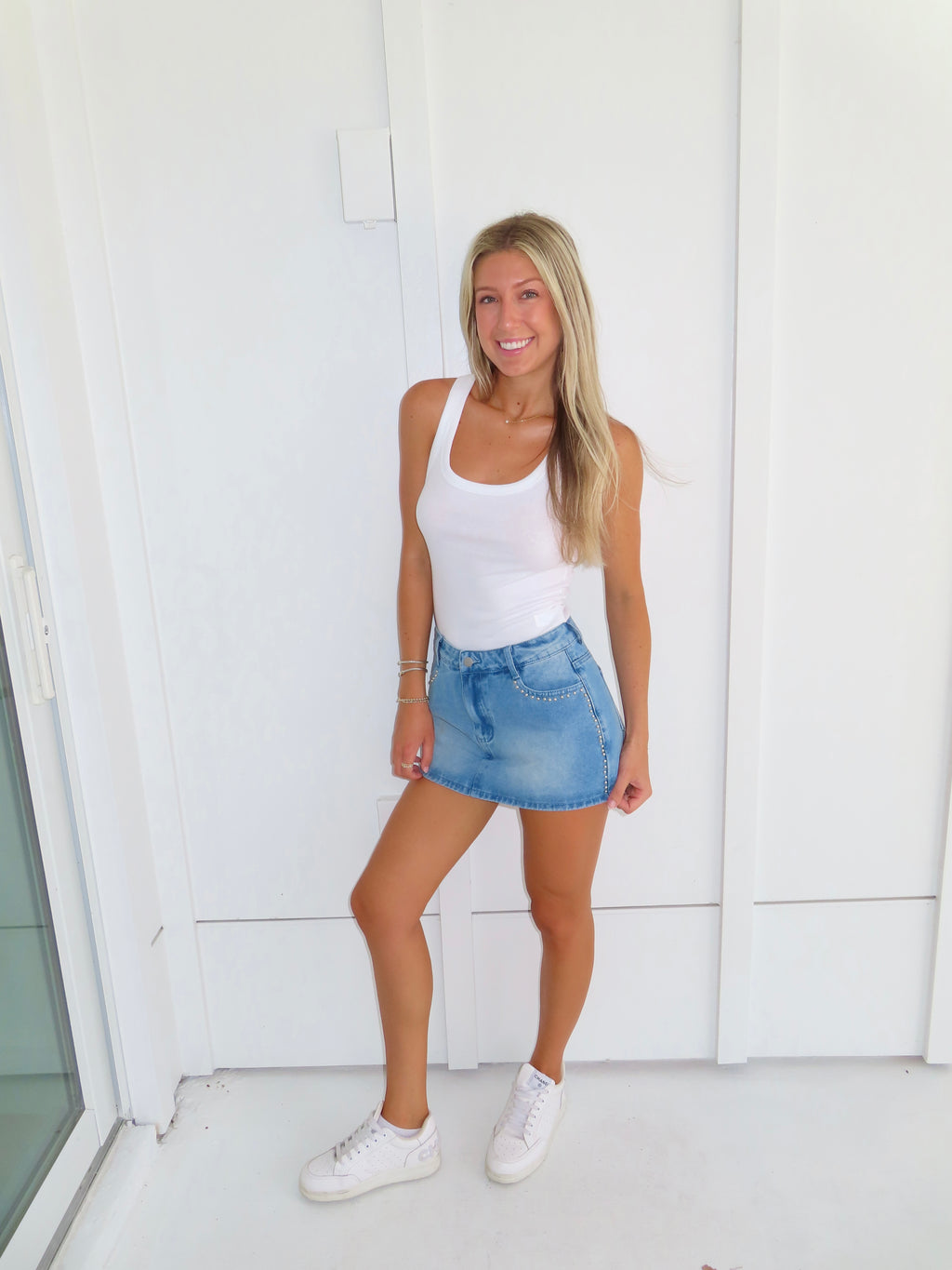 Woman wearing a white tank top and denim studded skirt with white sneakers standing against a white wall.