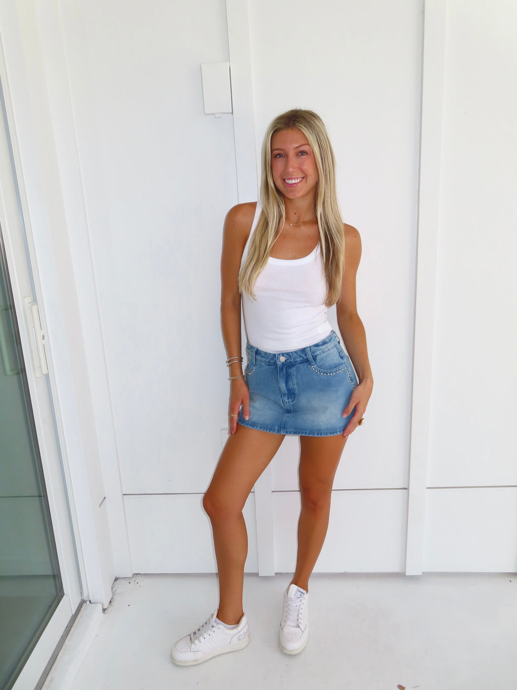 Woman wearing a white tank top and denim studded skirt with white sneakers standing against a white wall.