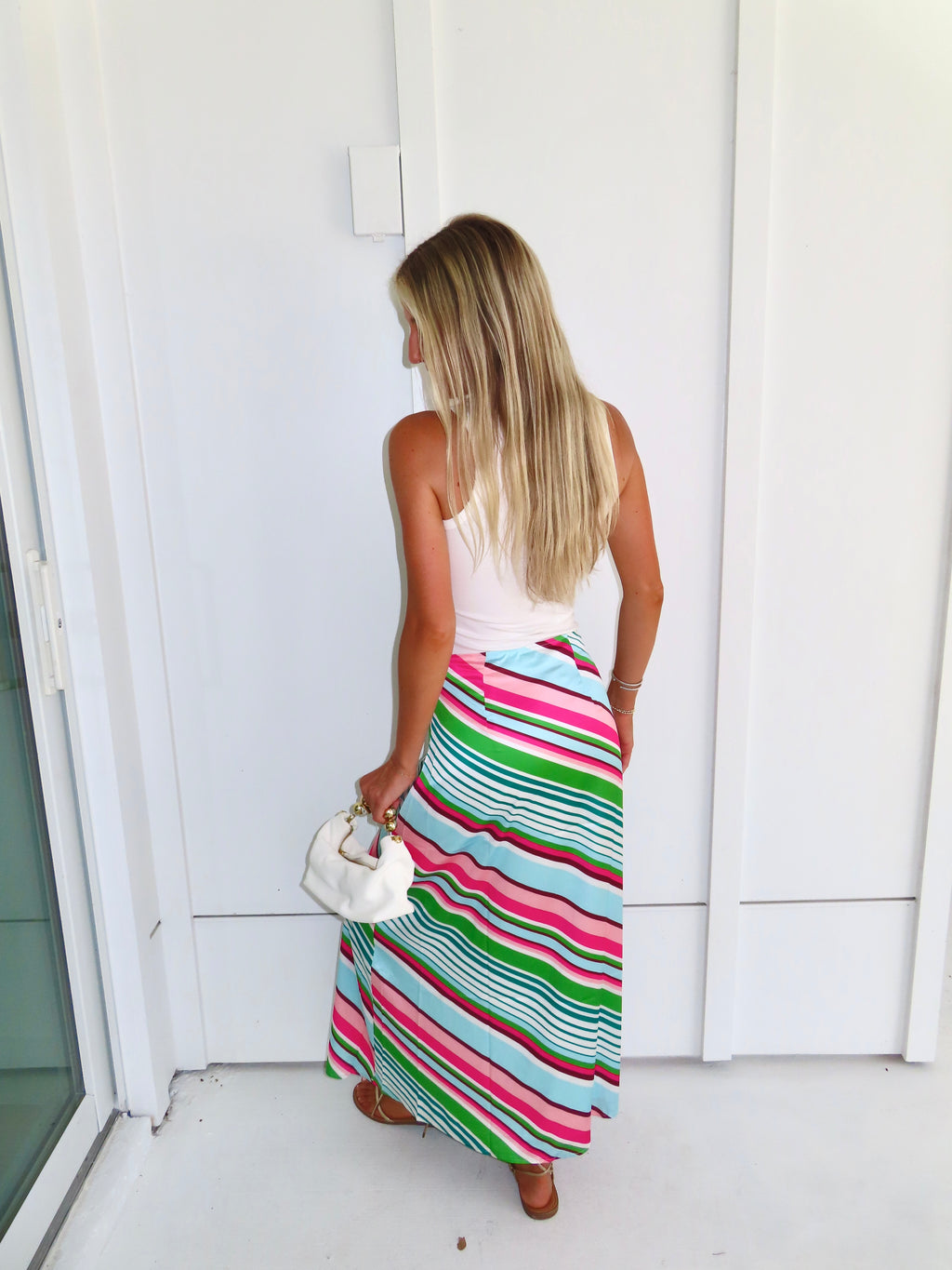 Woman wearing a white tank top and colorful striped skirt, holding a white purse and standing against a white wall.