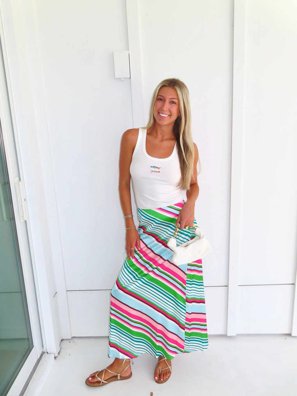Woman wearing a white tank top and colorful striped skirt, holding a white purse and standing against a white wall.