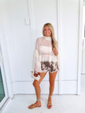 Woman wearing a cream ruffle top, denim cow printed shorts, and gold sandals holding a cream purse against a white wall.