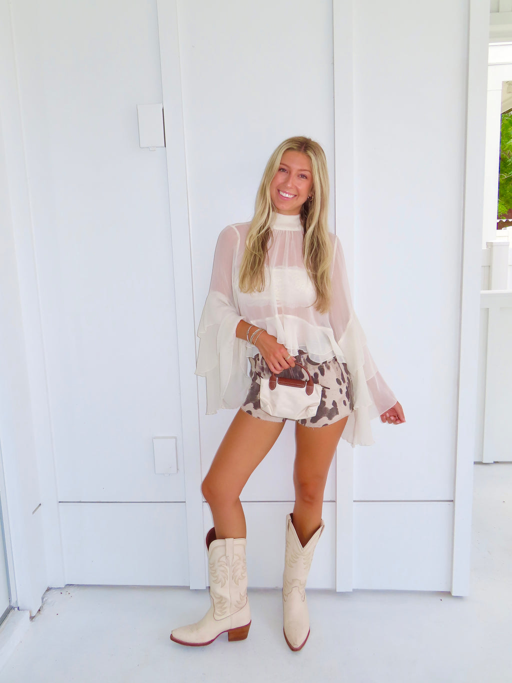 Woman wearing a cream ruffle top, denim cow printed shorts, and cream cowboy boots holding a cream purse against a white wall.