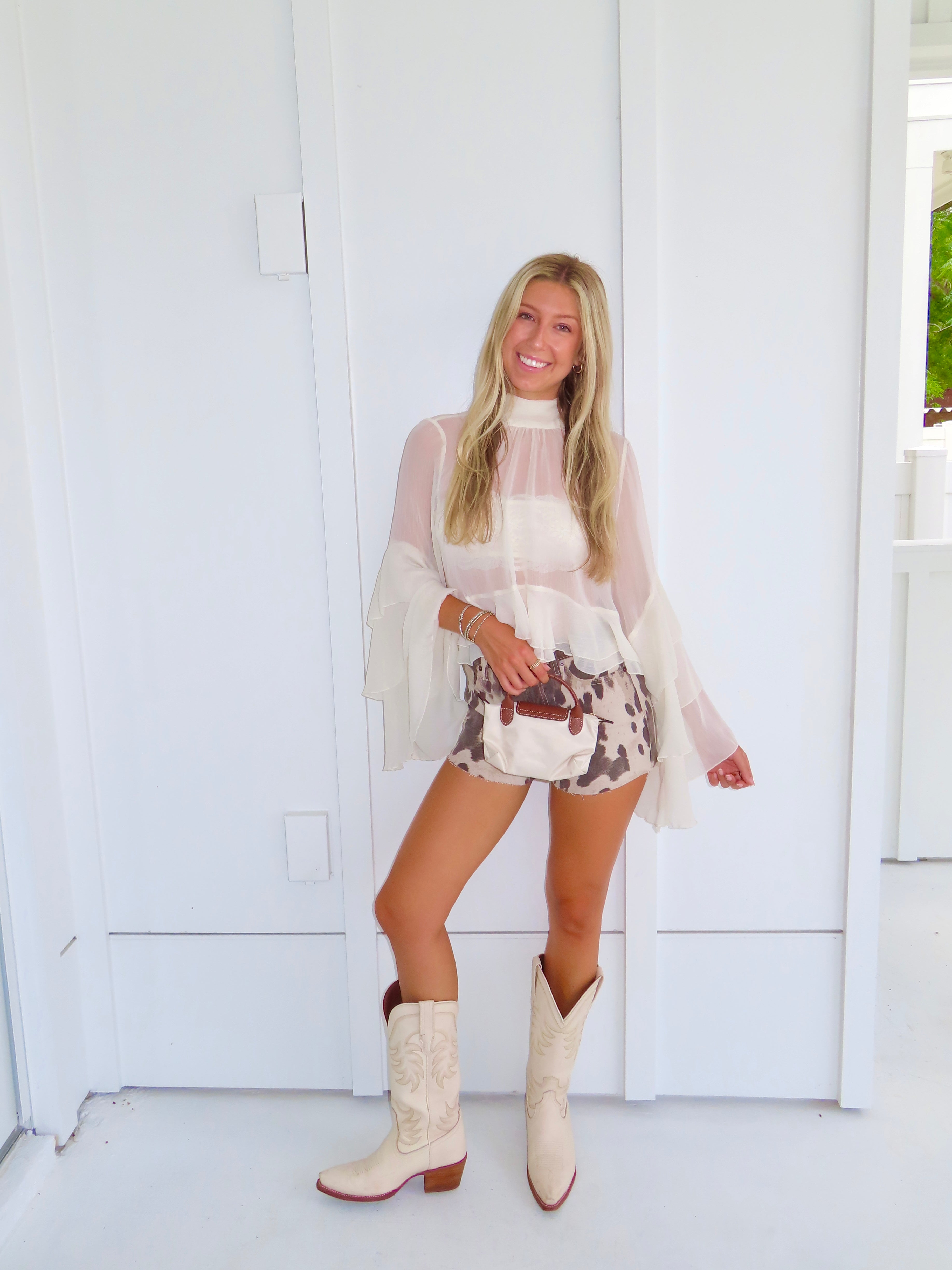 Woman wearing a cream ruffle top, denim cow printed shorts, and cream cowboy boots holding a cream purse against a white wall.
