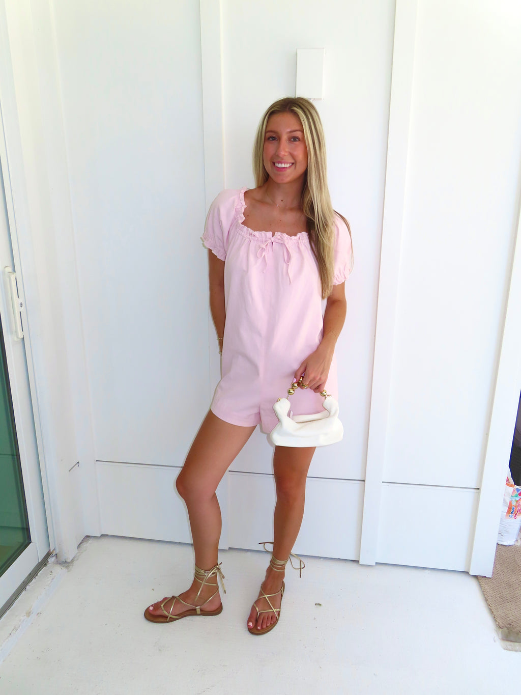 Woman wearing a pink romper and gold sandals and holding a white purse standing against a white wall.