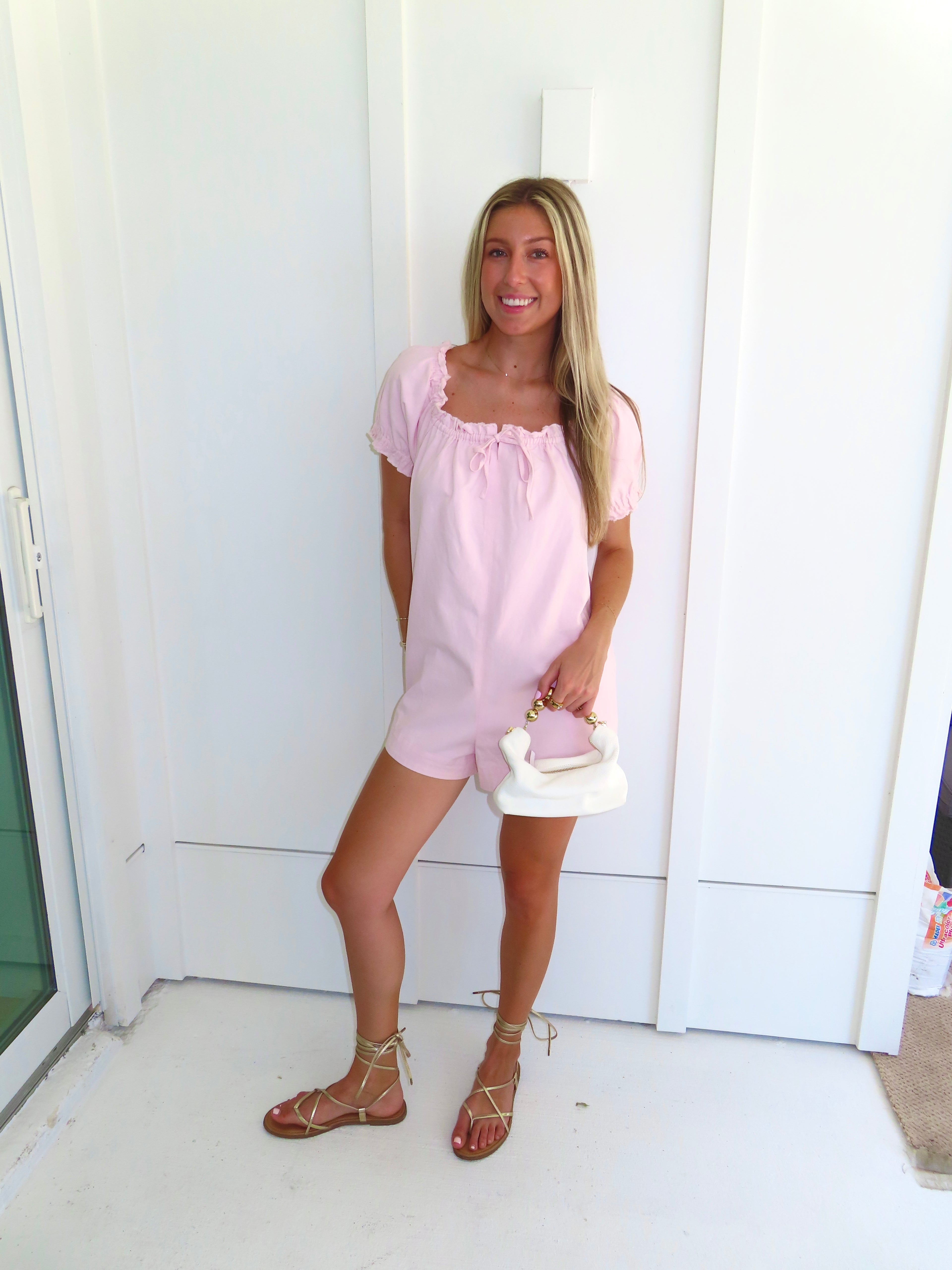 Woman wearing a pink romper and gold sandals and holding a white purse standing against a white wall.