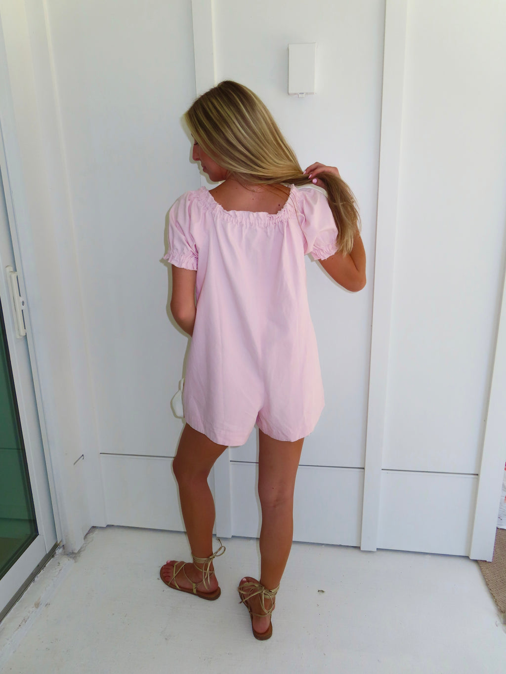Woman wearing a pink romper and gold sandals standing against a white wall.
