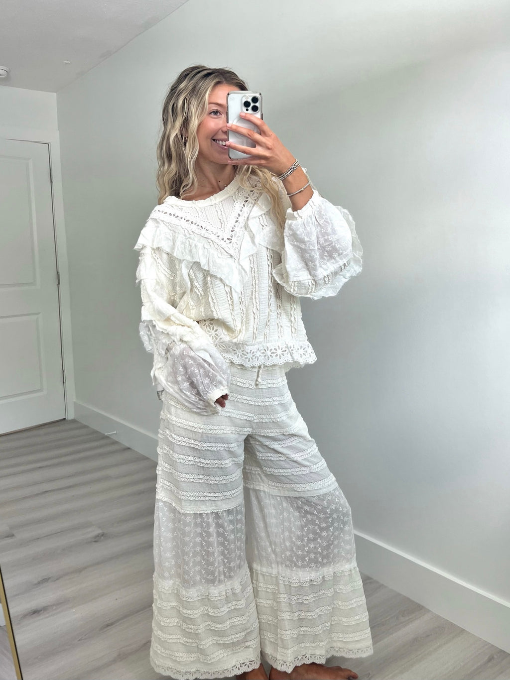 Woman taking a mirror selfie in a room with white walls and light wood floor wearing a cream lace ruffle long sleeve and patching pants set