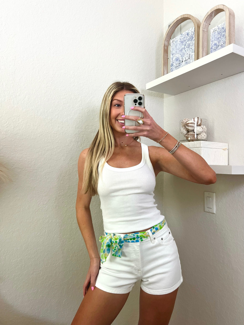 Woman taking a mirror selfie wearing a white tank top and white shorts with a colorful belt, holding a small handbag.