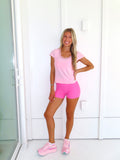 Woman wearing a pink tee and pink bikers shorts with pink sneakers standing against a white wall.
