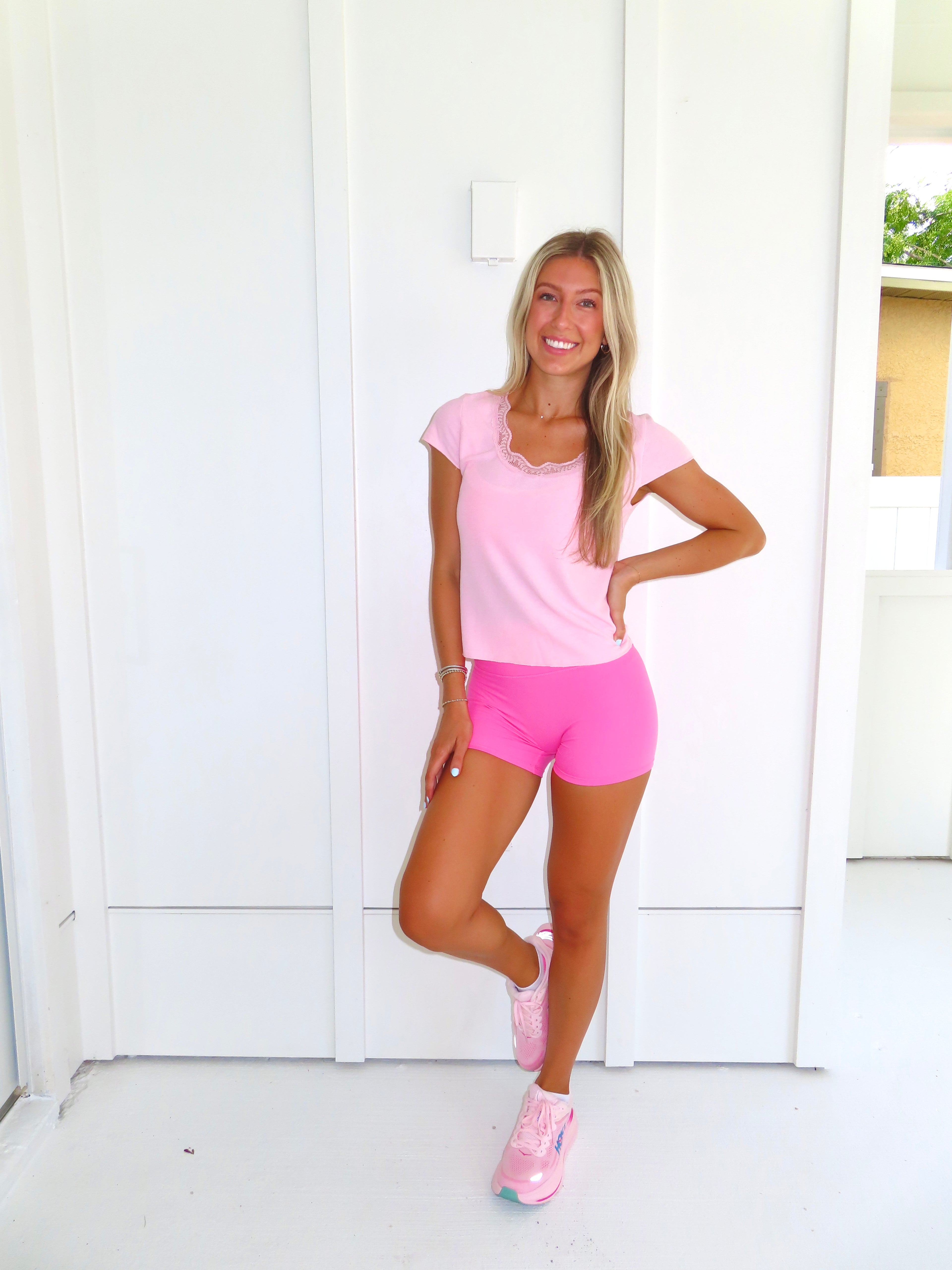 Woman wearing a pink tee and pink bikers shorts with pink sneakers standing against a white wall.